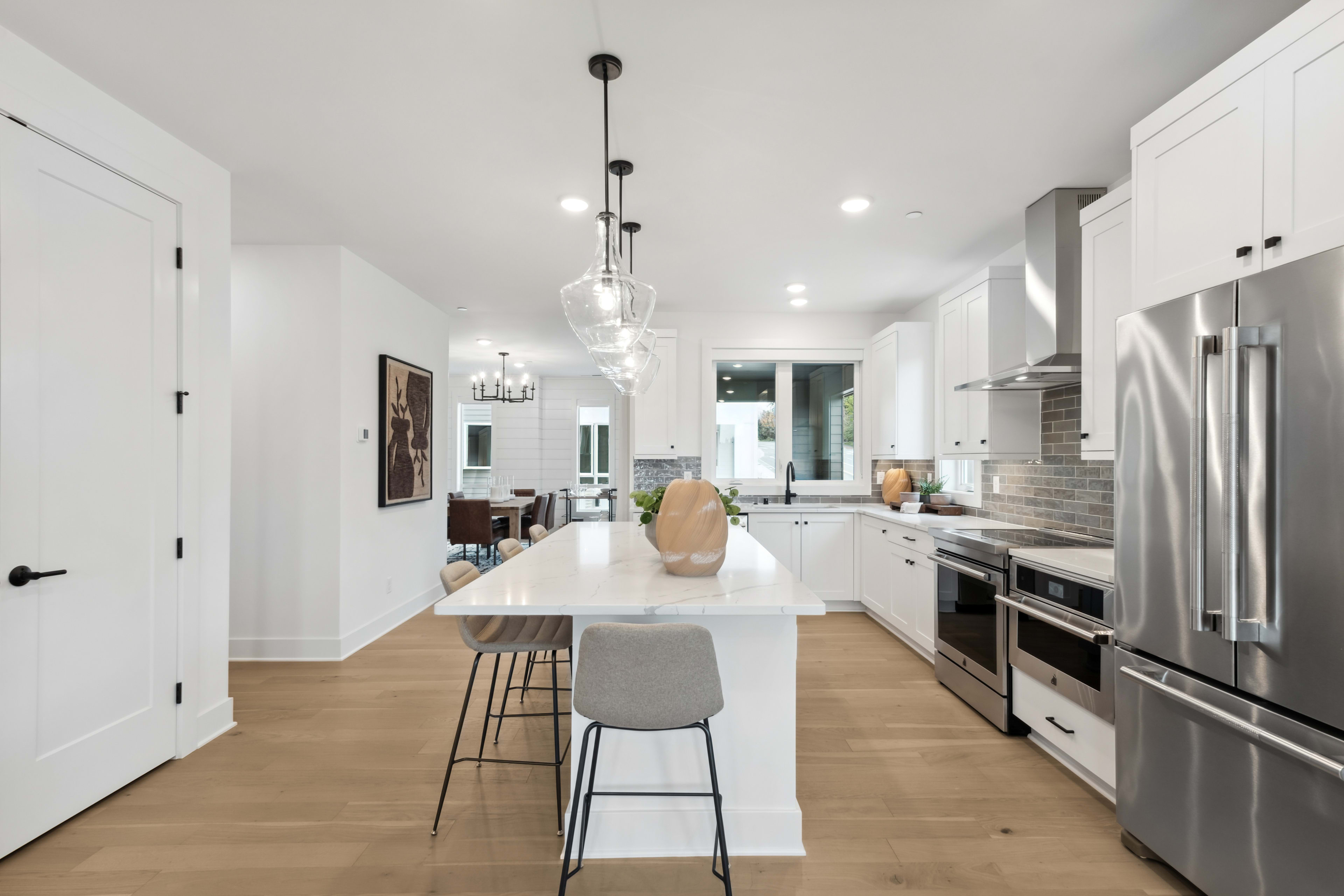 A modern, open-concept kitchen with a large white island, stainless steel appliances, and pendant lighting fixtures, set against a backdrop of white walls and hardwood floors.