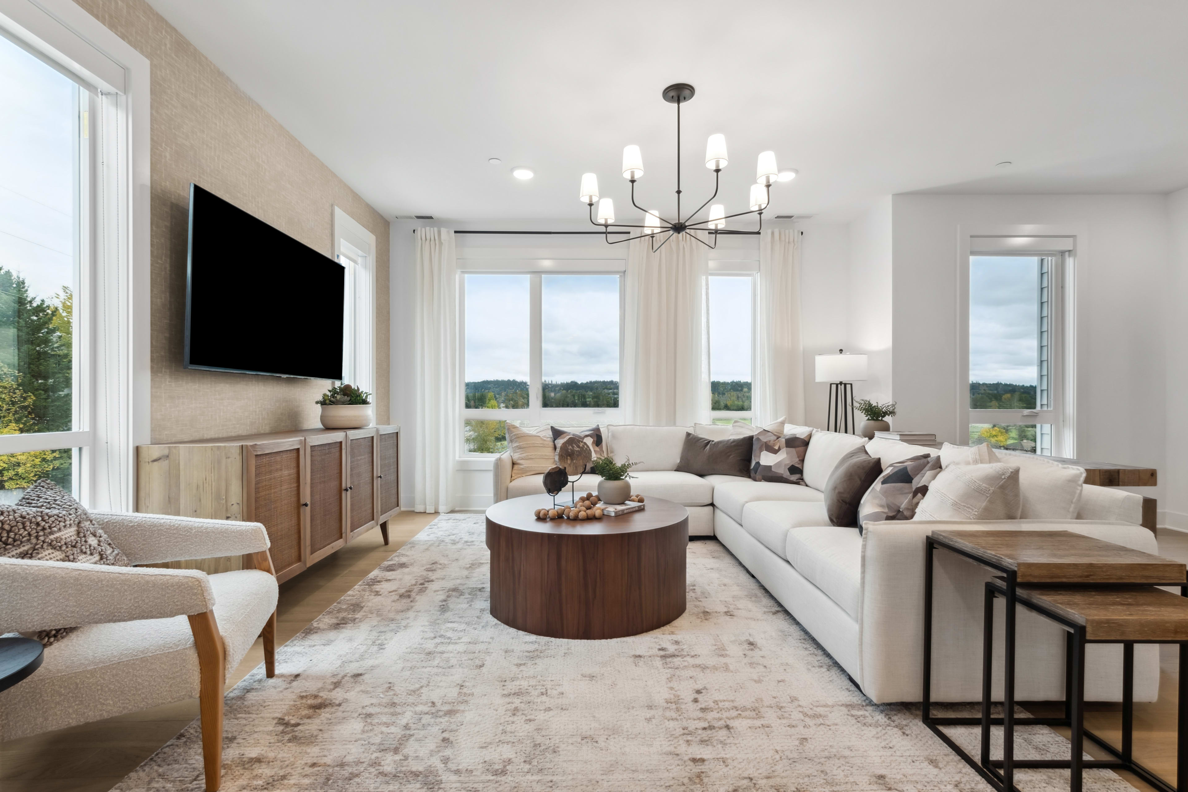 A spacious and modern living room with a large sectional sofa, a wooden coffee table, and a TV mounted on the wall, overlooking a scenic outdoor view through the large windows.