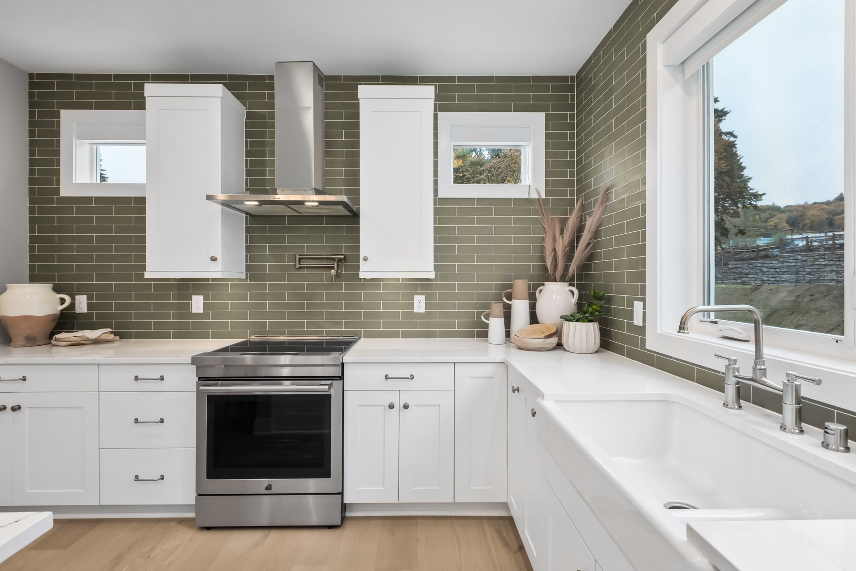 A modern, well-equipped kitchen with white cabinets, stainless steel appliances, and a backsplash of olive-green tiles, set against a scenic outdoor view through the window.