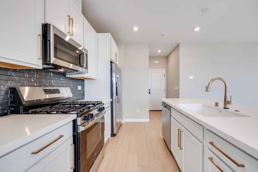 A modern, well-equipped kitchen with white cabinets, stainless steel appliances, and a tiled backsplash, leading into a hallway with a light-colored floor.