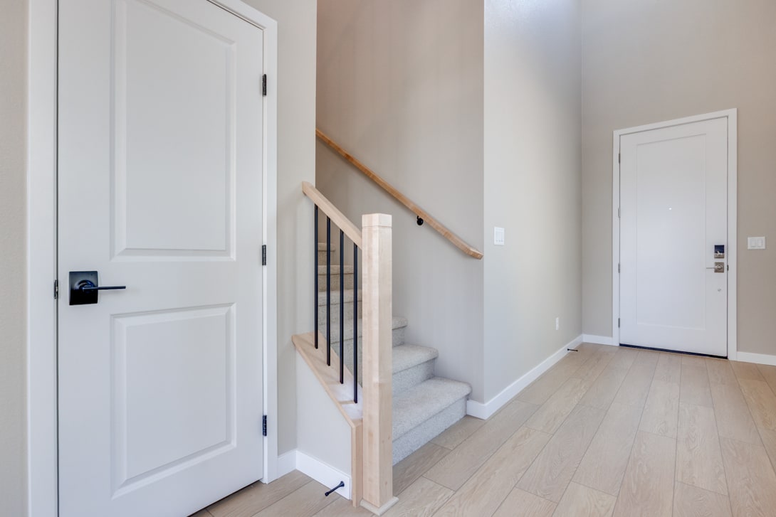 A bright and airy hallway with white doors, a wooden staircase, and a hardwood floor leading to another doorway.
