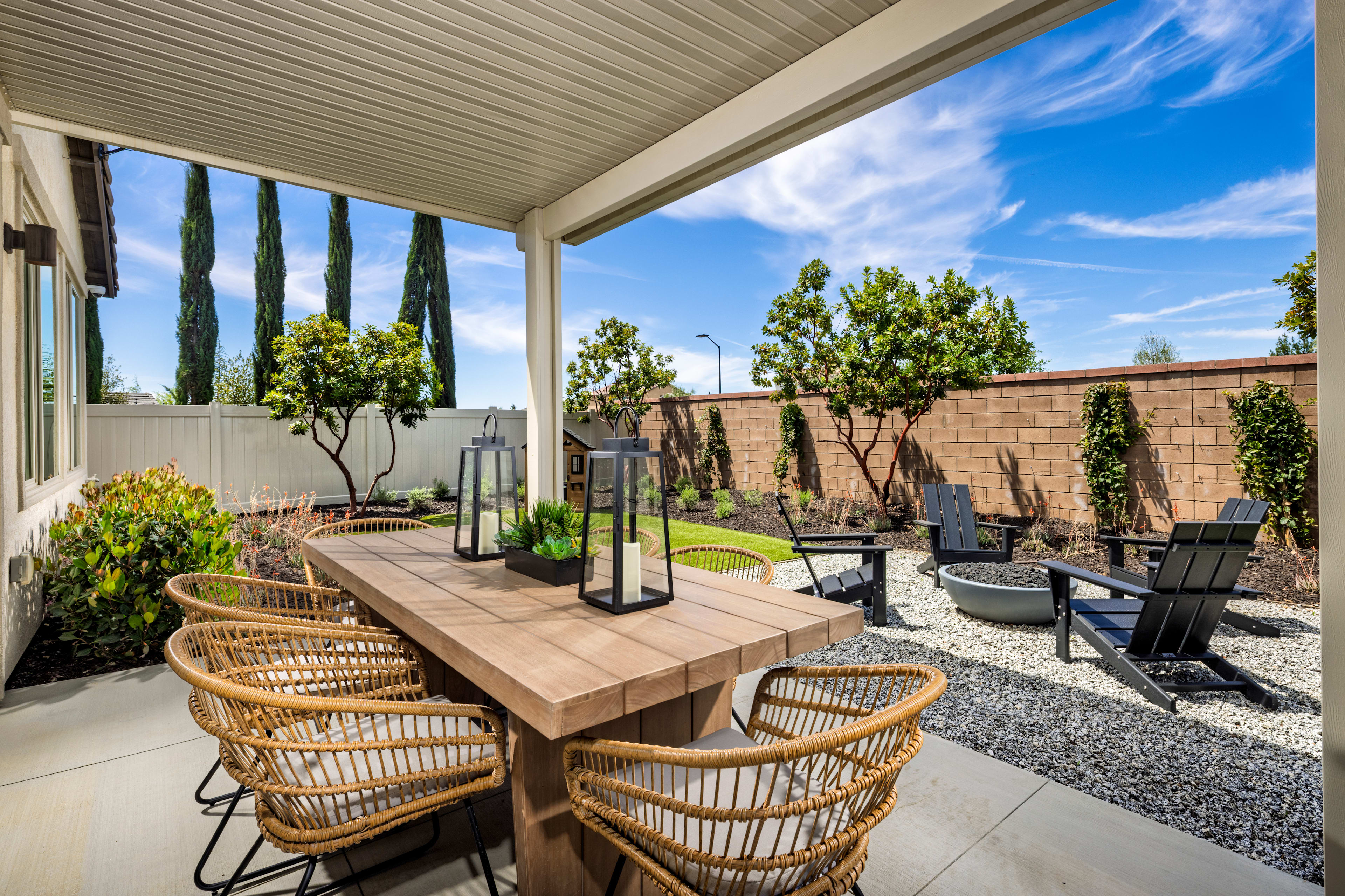 A cozy outdoor patio with a wooden table and wicker chairs, surrounded by lush greenery and a clear blue sky.