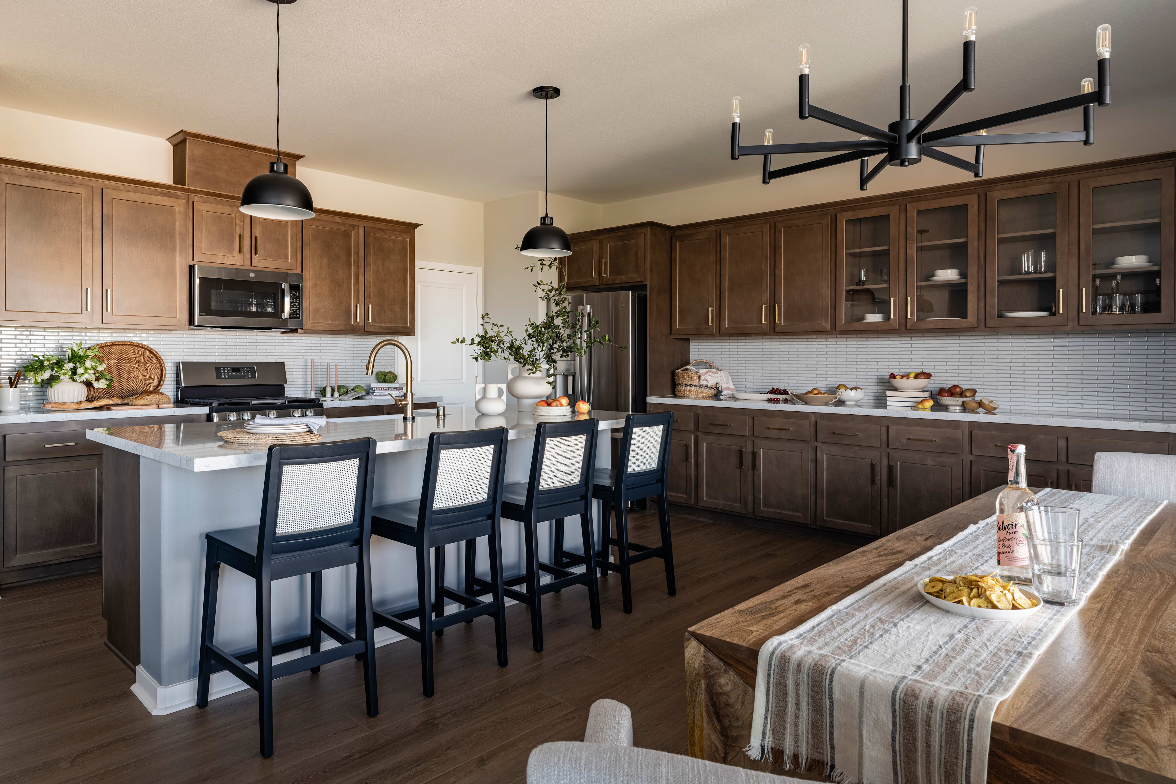 A modern and spacious kitchen with wooden cabinets, a large island with bar stools, and a chandelier overhead, set against a backdrop of a dining area with a table and chairs.
