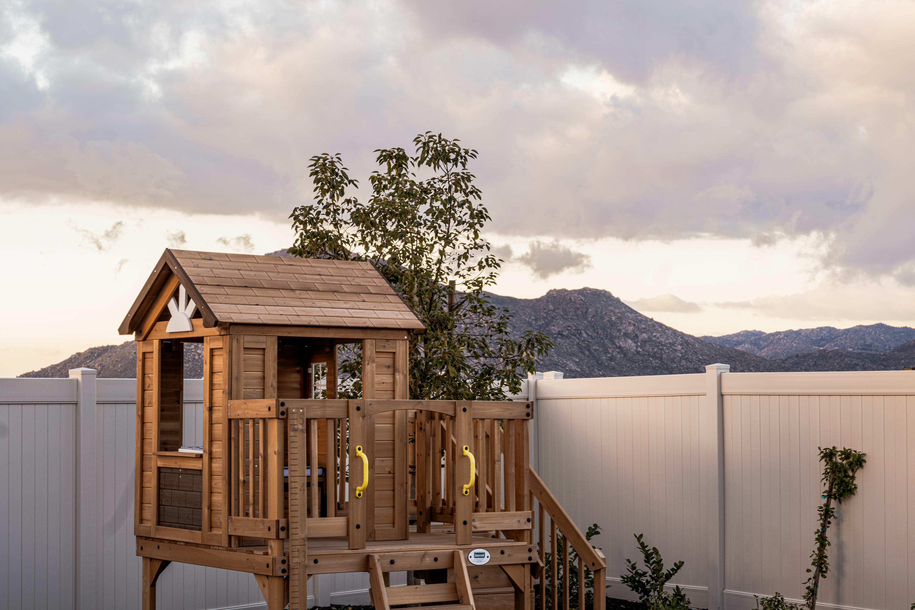 A wooden playhouse with a tree growing next to it, set against a backdrop of cloudy skies and a mountainous landscape.