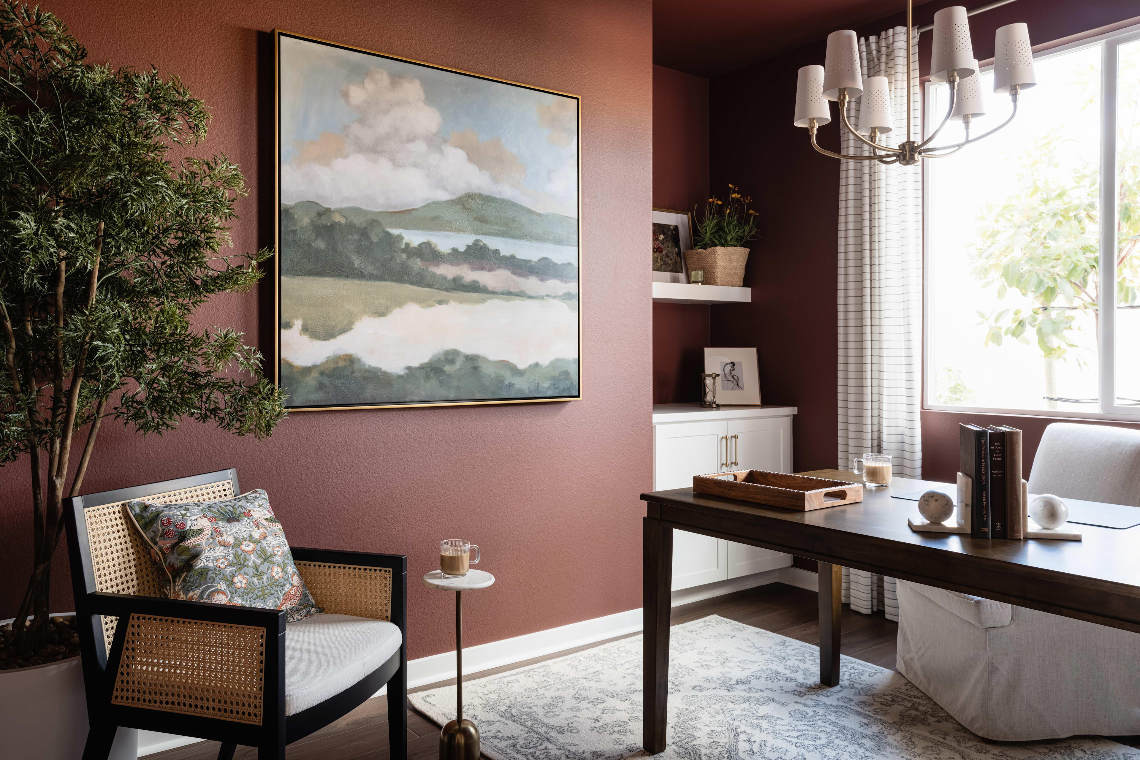 A cozy and inviting home office space with a large landscape painting on the wall, a wicker chair, and a wooden desk with various decorative elements.