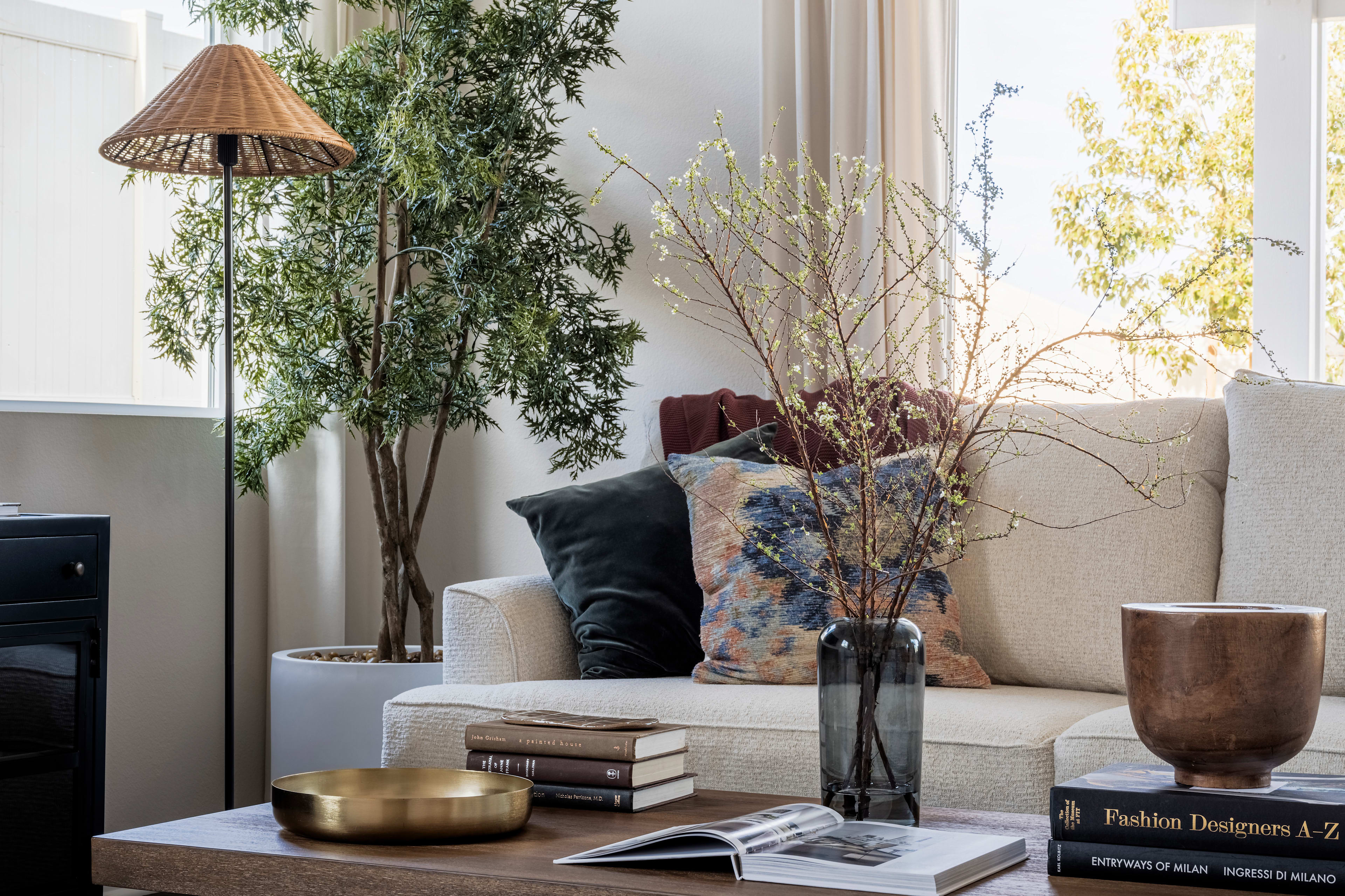 A cozy and inviting living room with a large wooden coffee table, a potted plant, and a vase of flowers, set against a backdrop of large windows and curtains.
