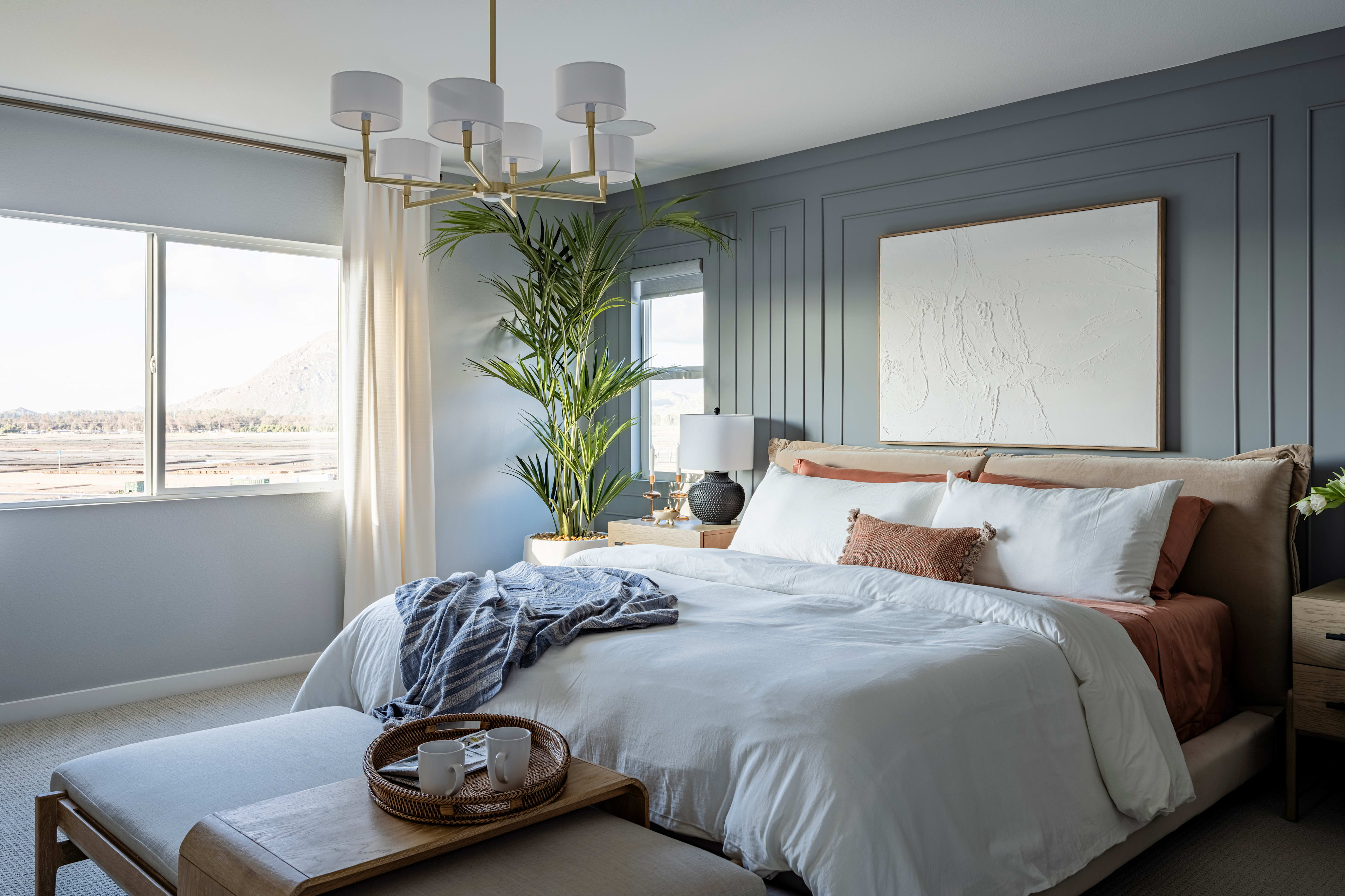 A cozy and inviting bedroom with a large bed, neutral-toned bedding, and a wooden bench at the foot of the bed. The room features gray walls, a chandelier, and a large window overlooking a scenic view.