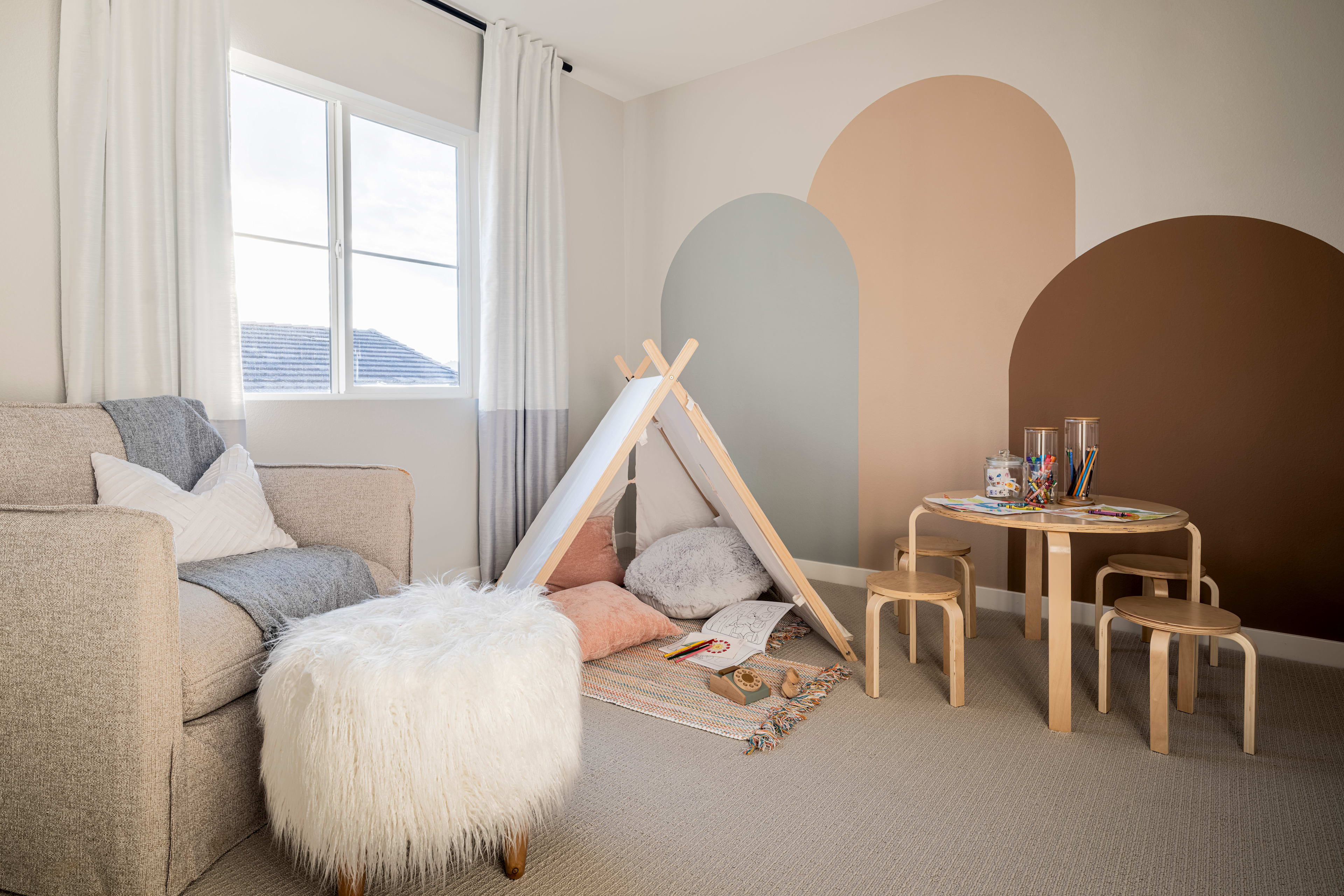 A cozy and inviting living space with a teepee-style play area, plush seating, and a wooden table and stools in the background, set against a neutral-toned wall with geometric shapes.