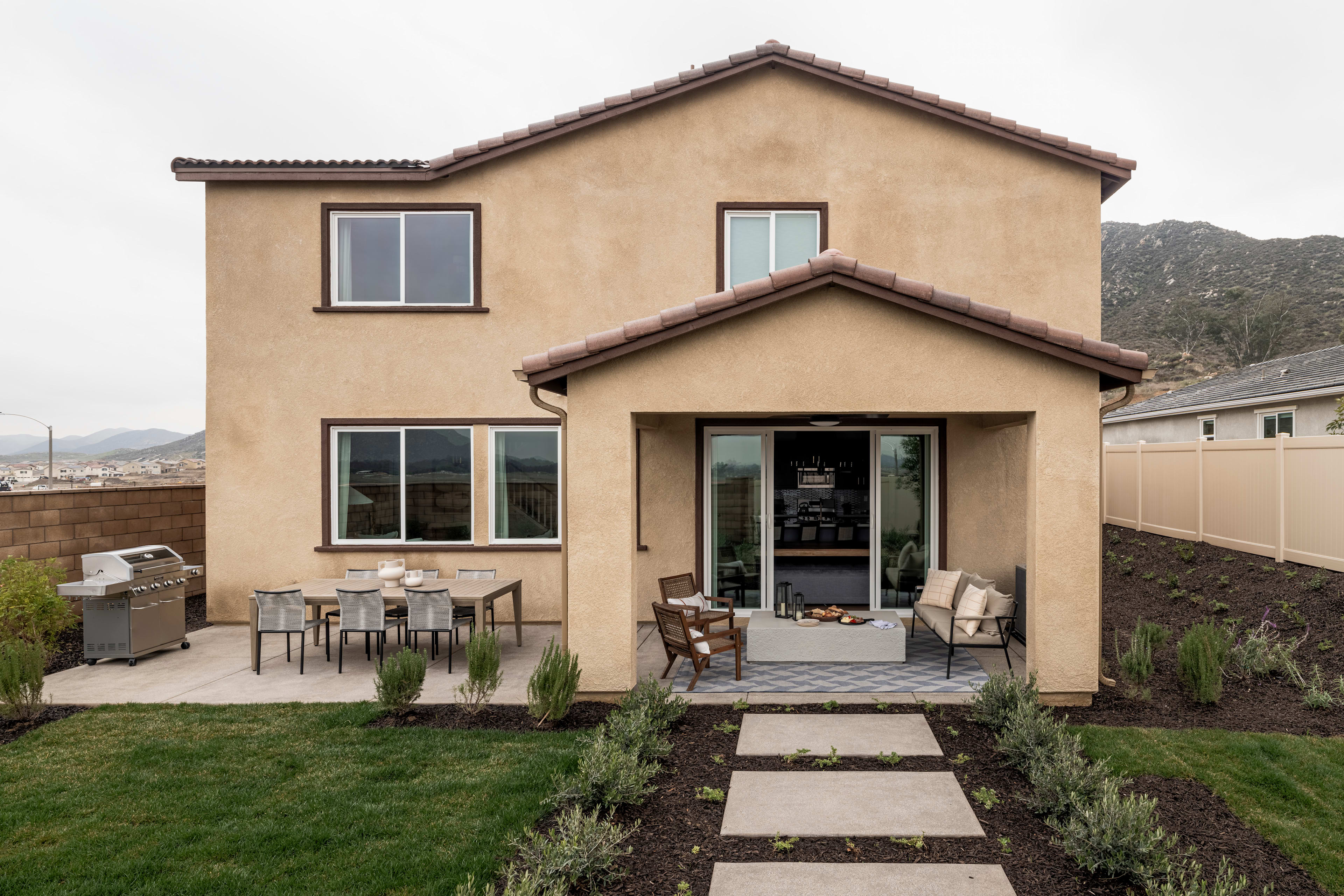 A two-story stucco house with a tiled roof sits on a grassy lawn, surrounded by a fence and landscaping. In the foreground, a paved walkway leads to the front porch, which features outdoor furniture and decor.