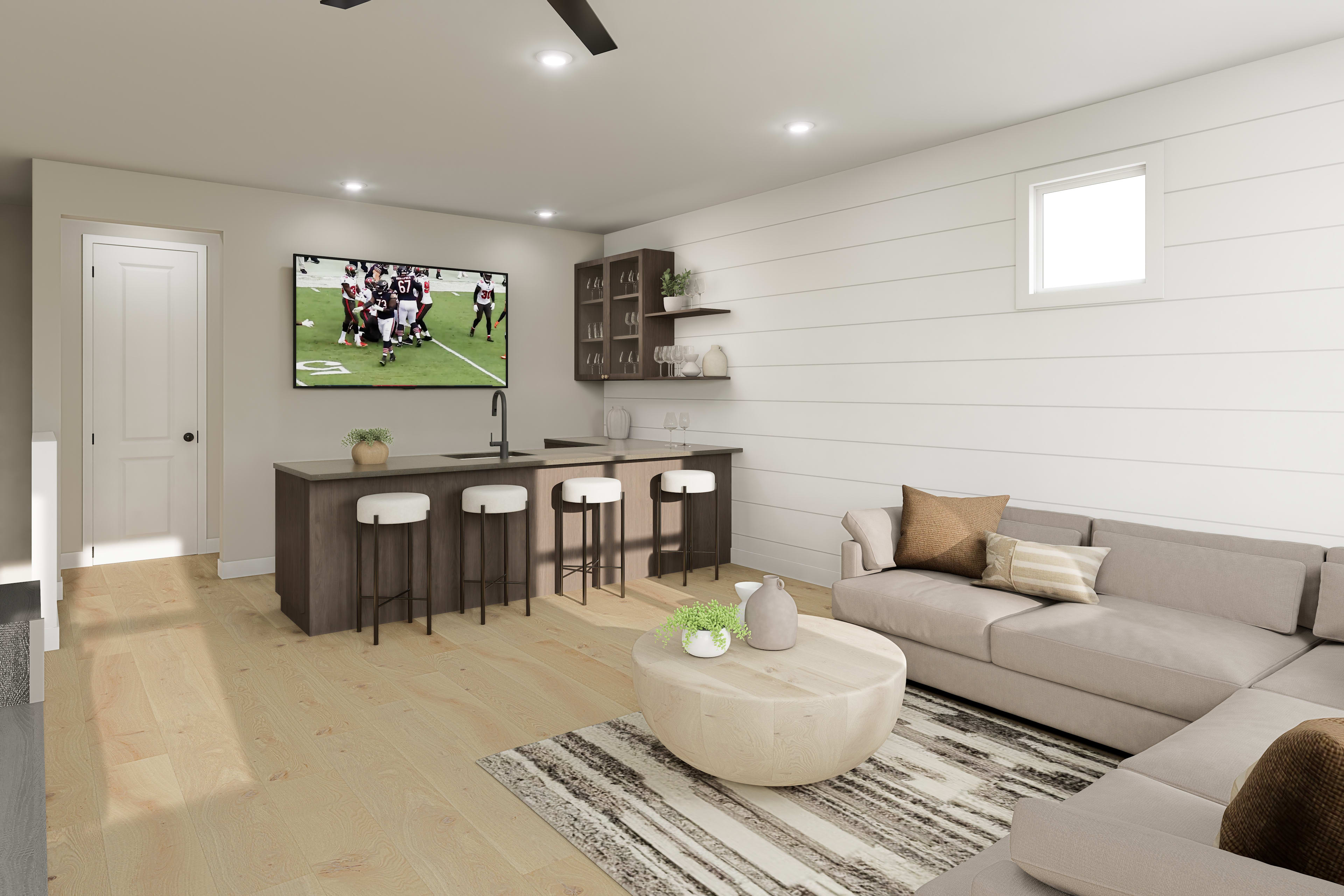 A modern and cozy living room with a large TV displaying a sports game, a gray sectional sofa, a round ottoman, and a wooden bar area with stools.