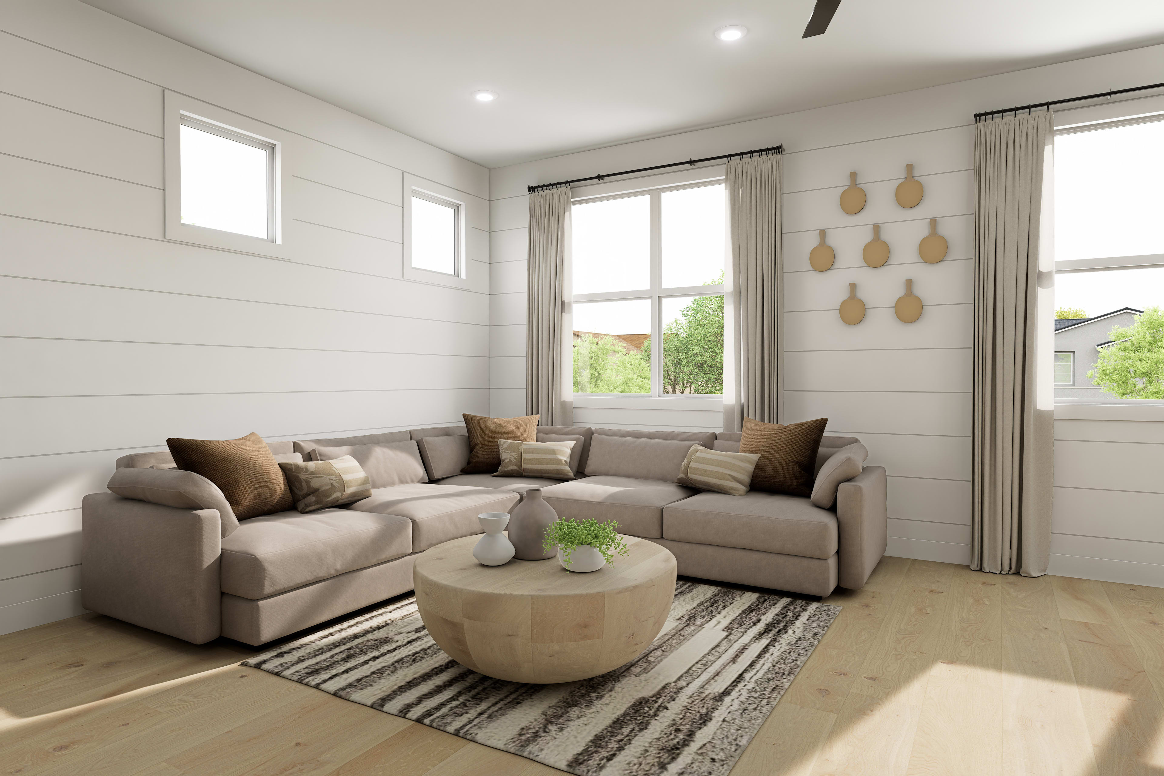 A cozy and inviting living room with a comfortable sectional sofa, a round wooden coffee table, and decorative elements such as wall art and potted plants, set against a backdrop of white shiplap walls and large windows overlooking a lush outdoor scene.