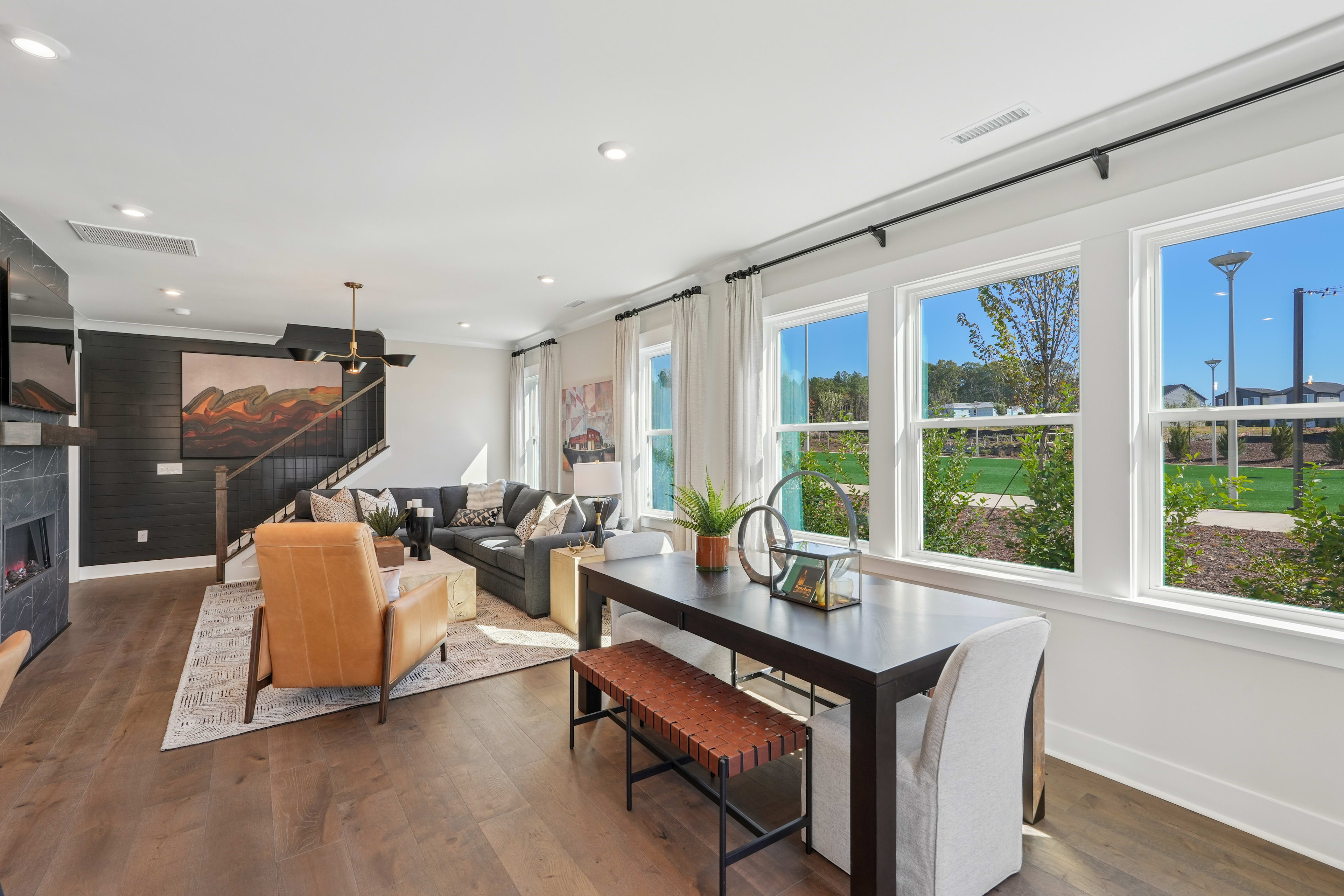 A modern and spacious living room with large windows overlooking a lush outdoor landscape, featuring a sleek black table, comfortable seating, and a warm, inviting atmosphere.