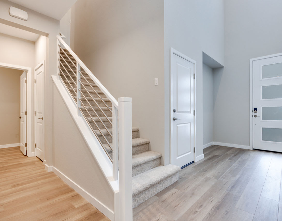 A bright and spacious entryway with a staircase leading upstairs, hardwood floors, and a white door in the background.