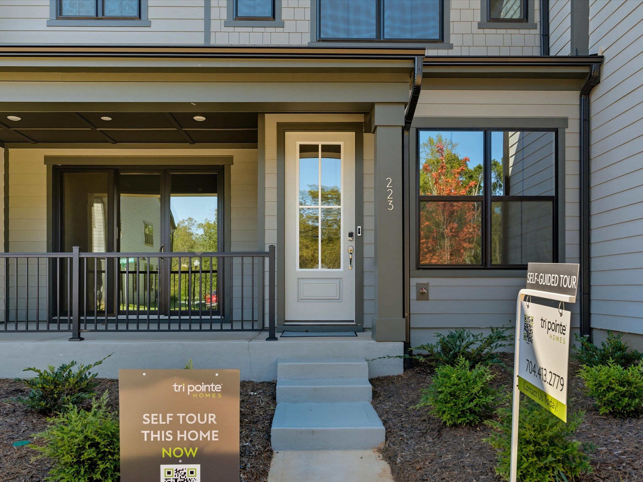 A modern two-story residential building with a covered porch, surrounded by landscaped greenery and a "For Sale" sign in the foreground.