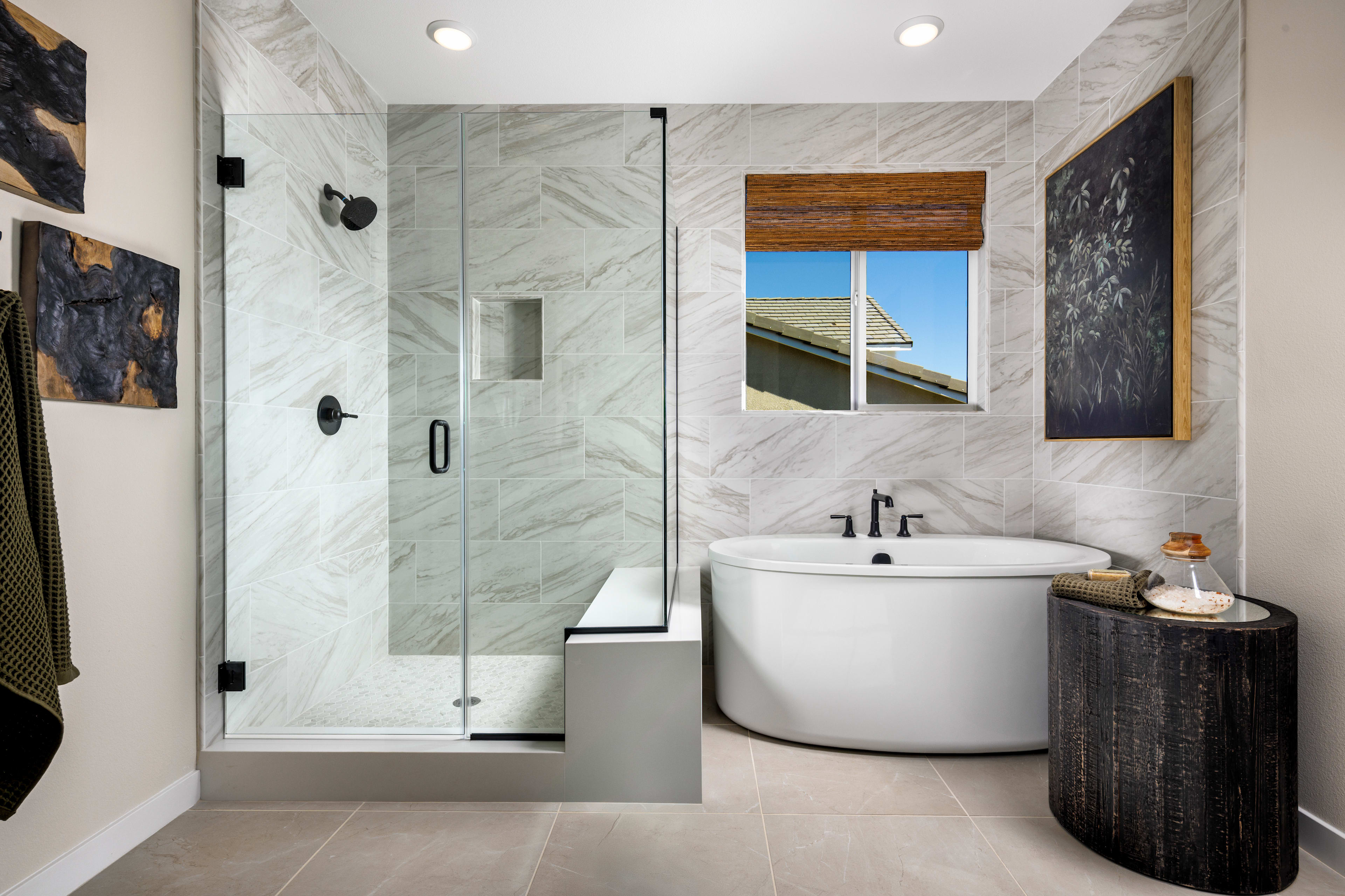 A modern and spacious bathroom with a freestanding bathtub, glass shower enclosure, and natural stone tiles on the walls and floor. The room features a large window providing natural light and a view of the exterior.