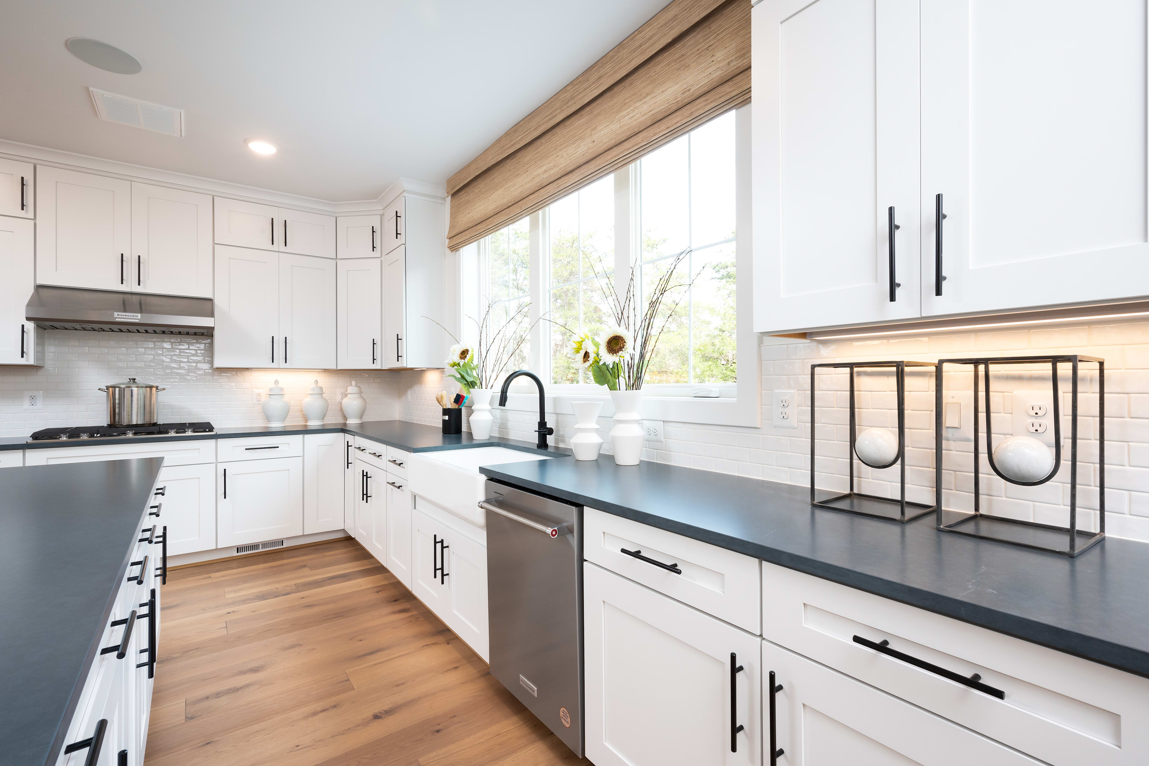 A modern, well-designed kitchen with white cabinets, wooden accents, and a sleek black countertop, featuring a window that provides natural lighting and a view of the outdoors.