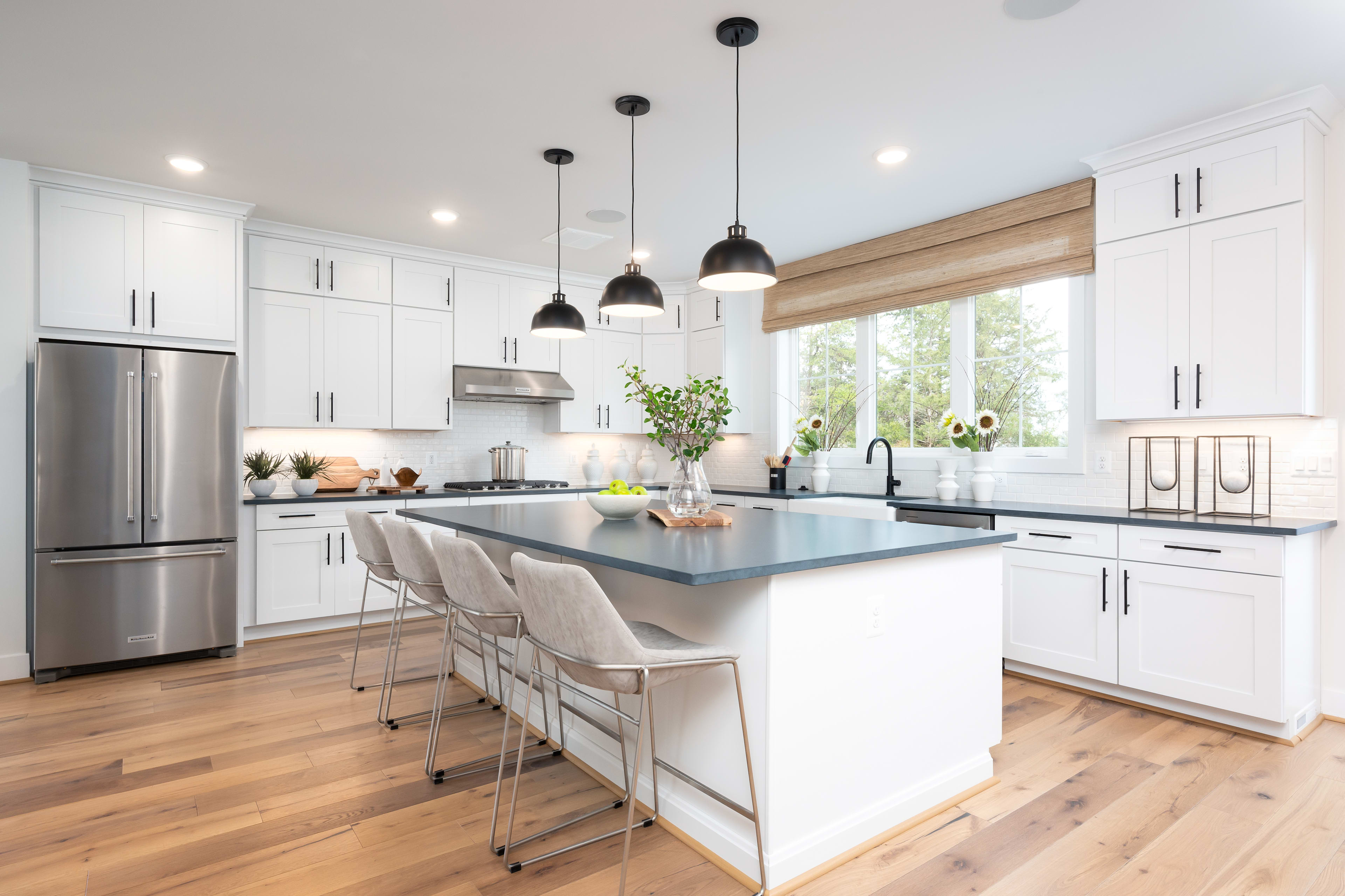 A modern, bright kitchen with white cabinets, a large island with stools, and pendant lighting over the island. The kitchen has hardwood floors and large windows overlooking the outdoors.