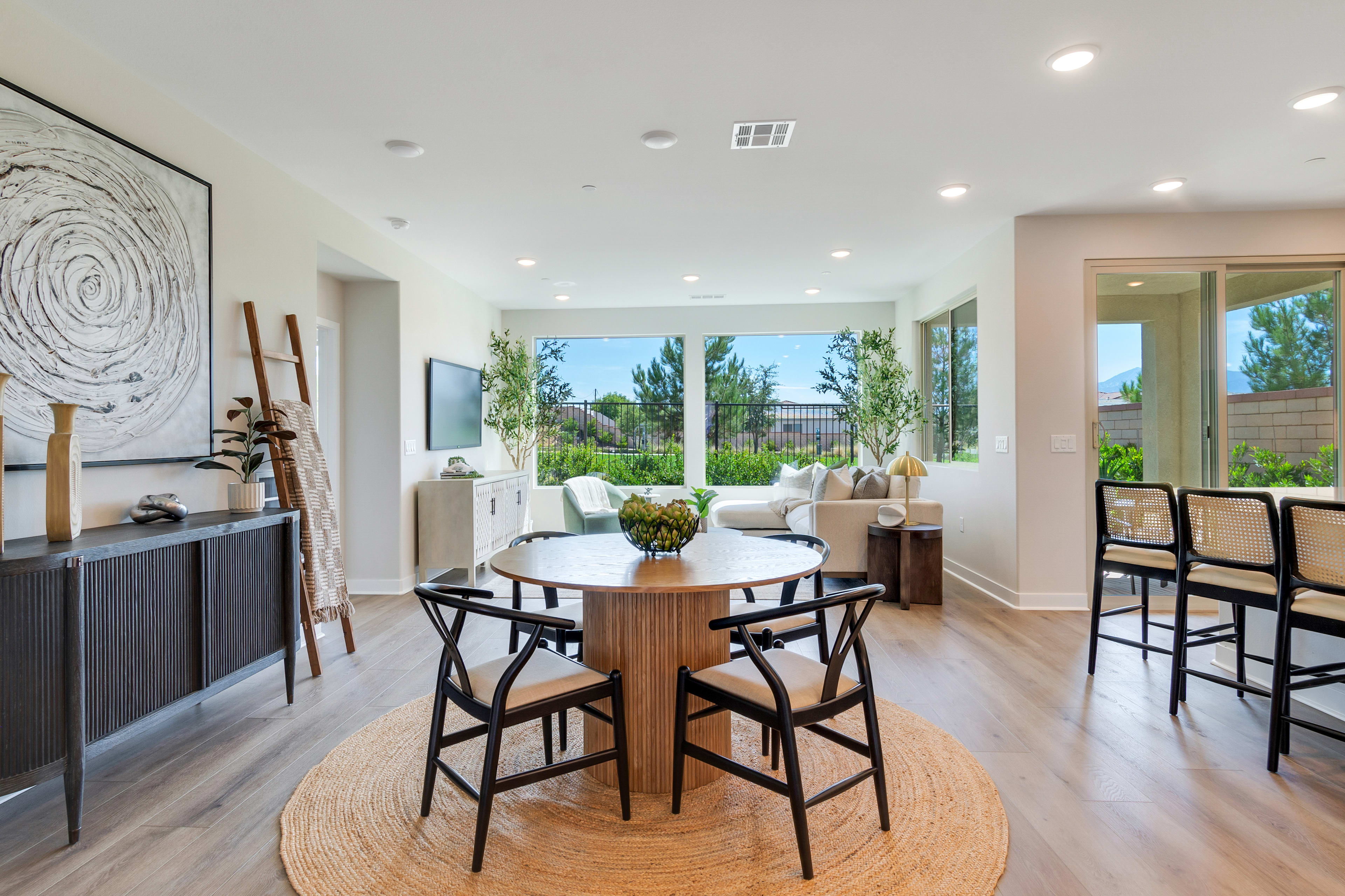 A modern and spacious dining area with a round wooden table and black bar stools, surrounded by large windows overlooking a lush outdoor landscape.
