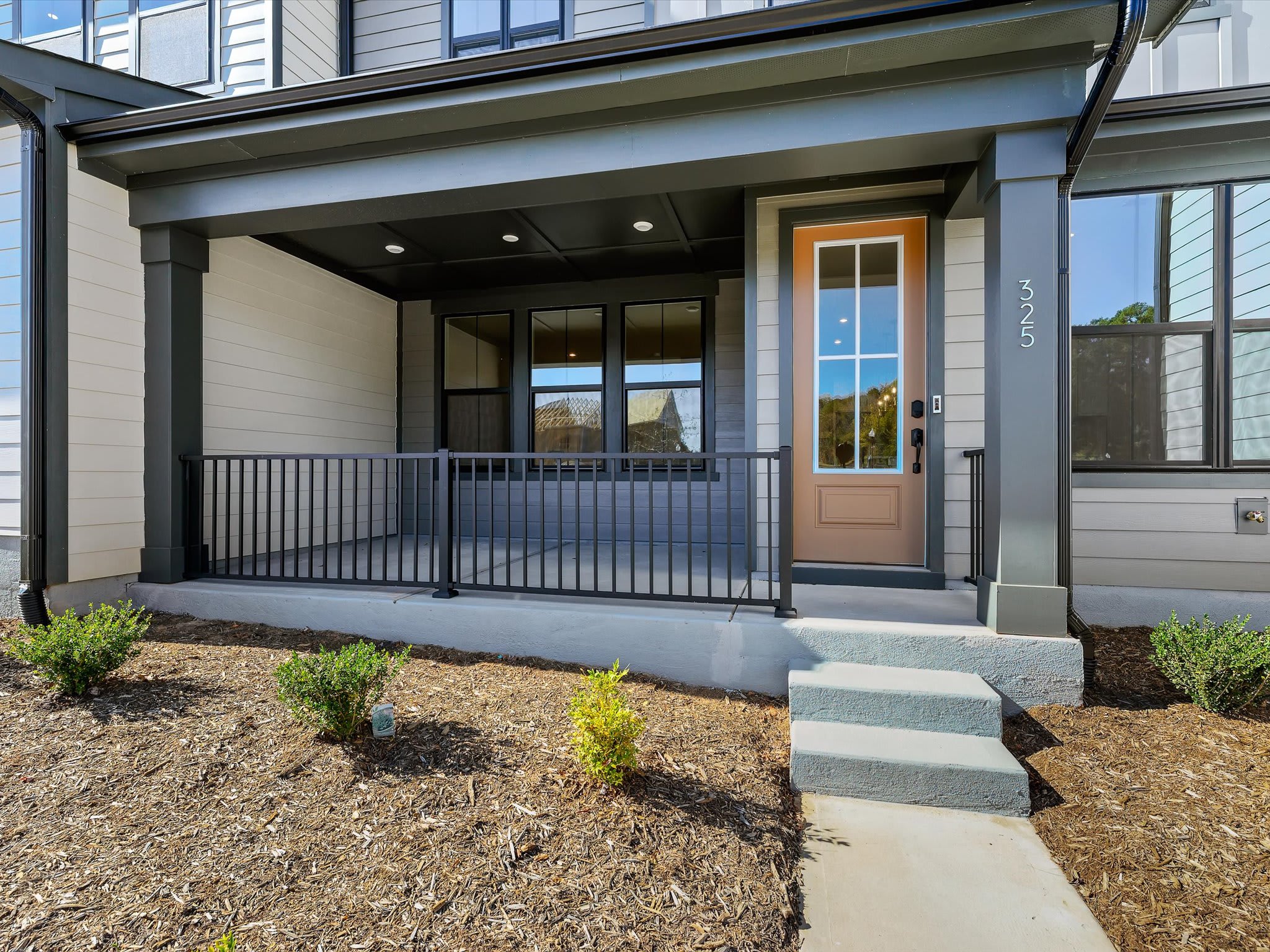The image shows a modern, two-story residential building with a covered porch entrance, surrounded by a landscaped yard with a concrete walkway leading to the front door.