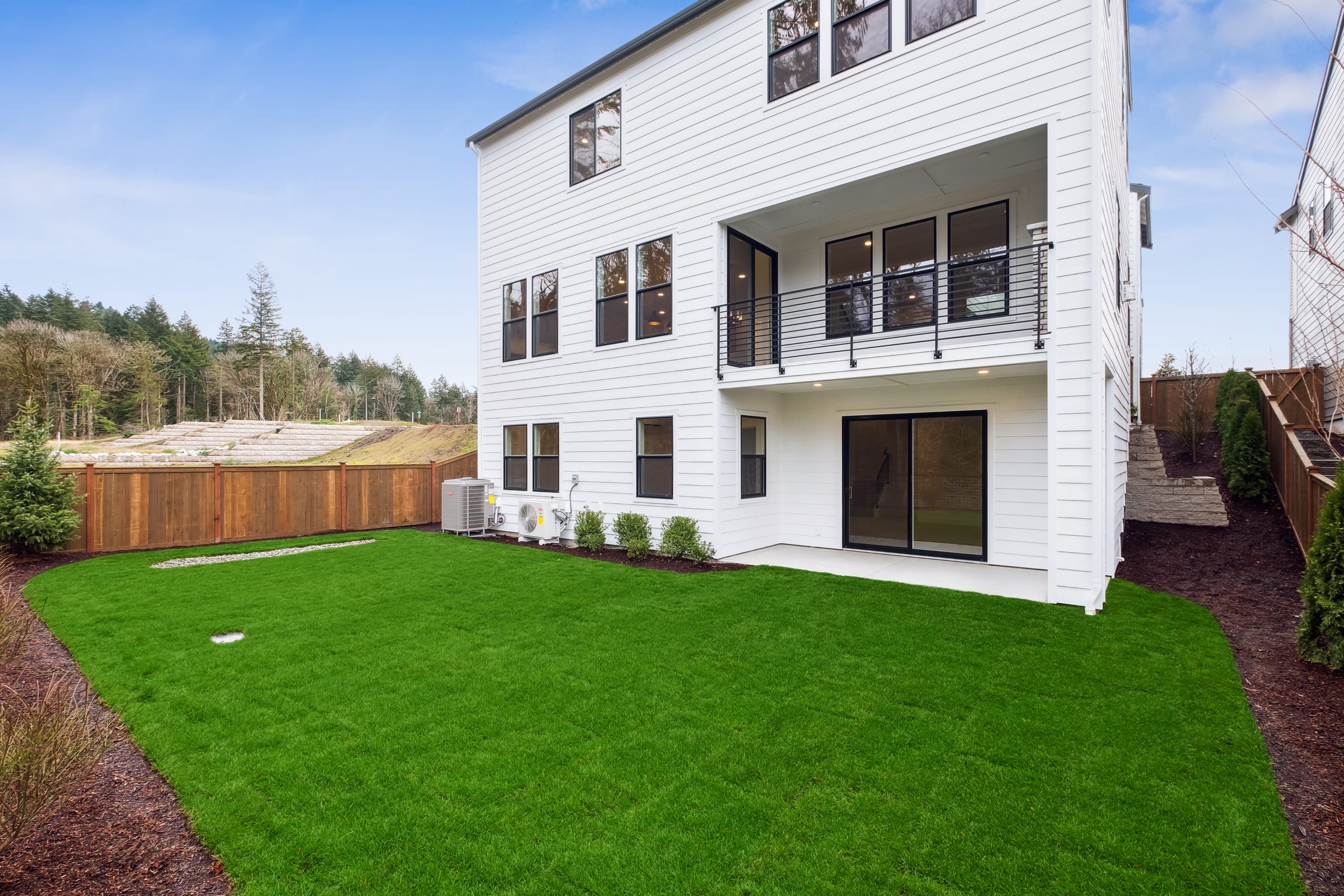 A modern, two-story white house with a well-manicured green lawn in the foreground, surrounded by a wooded area in the background.