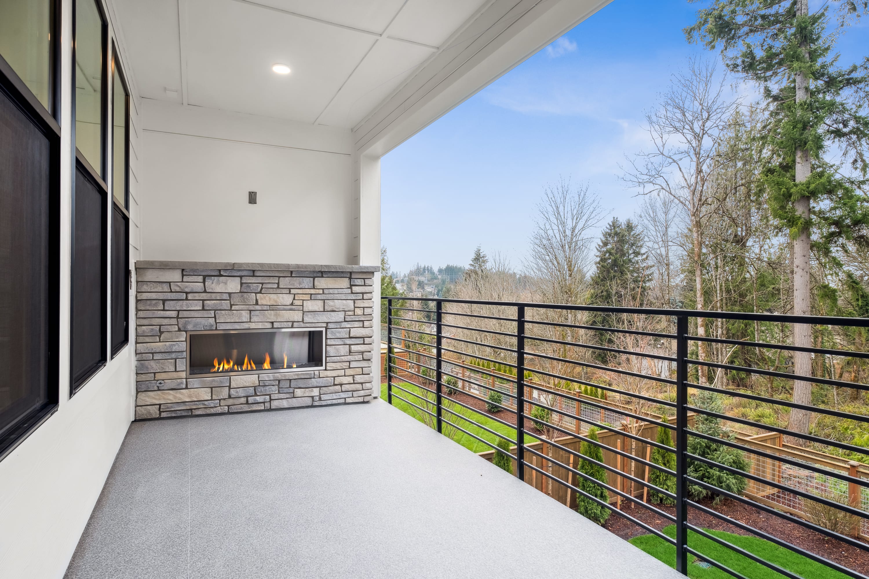 A modern outdoor patio with a fireplace, surrounded by a metal railing and overlooking a forested landscape.