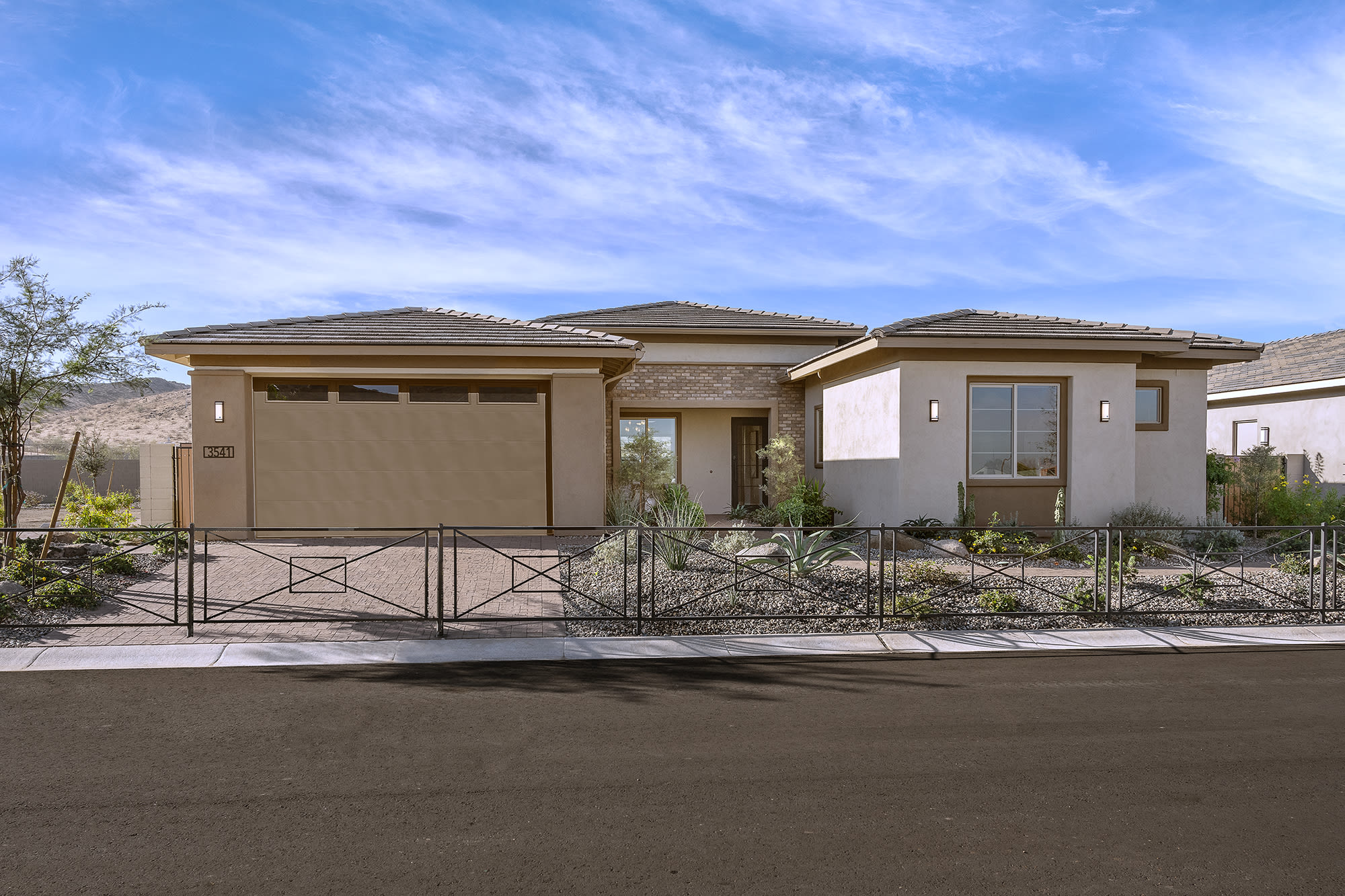 A single-story, beige-colored house with a tiled roof and a two-car garage stands in the foreground, surrounded by a paved driveway and a low metal fence. The background features a clear blue sky with wispy clouds.