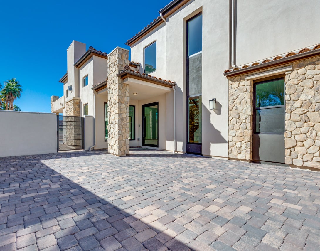 A modern, multi-story residential building with a stone facade, surrounded by a paved driveway and lush landscaping against a clear blue sky.