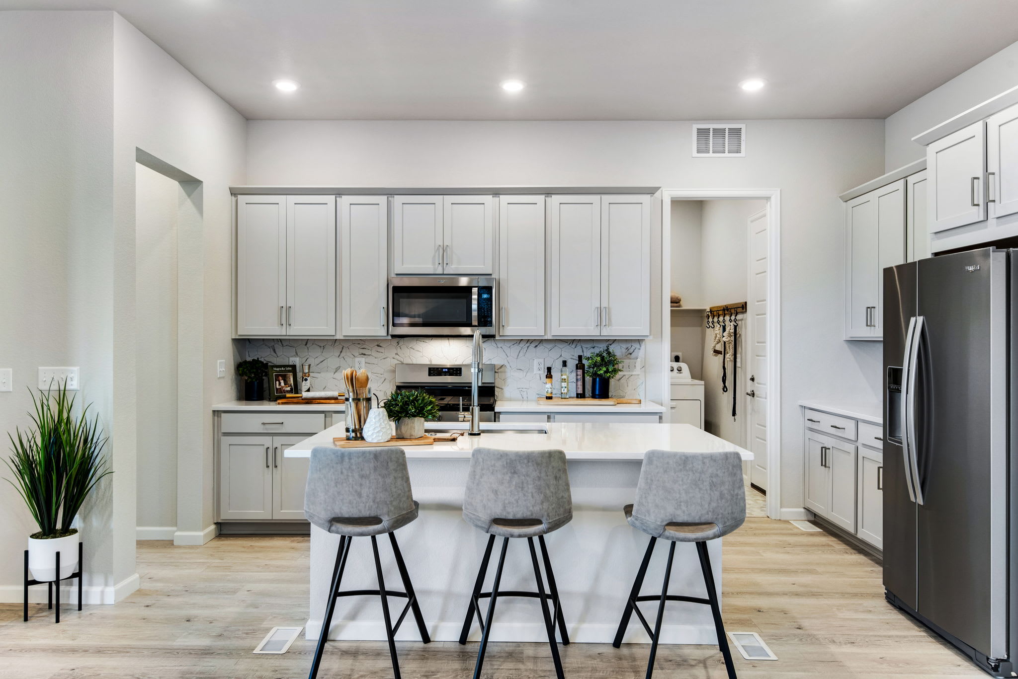 A modern, well-lit kitchen with white cabinets, stainless steel appliances, and a central island with three gray bar stools.