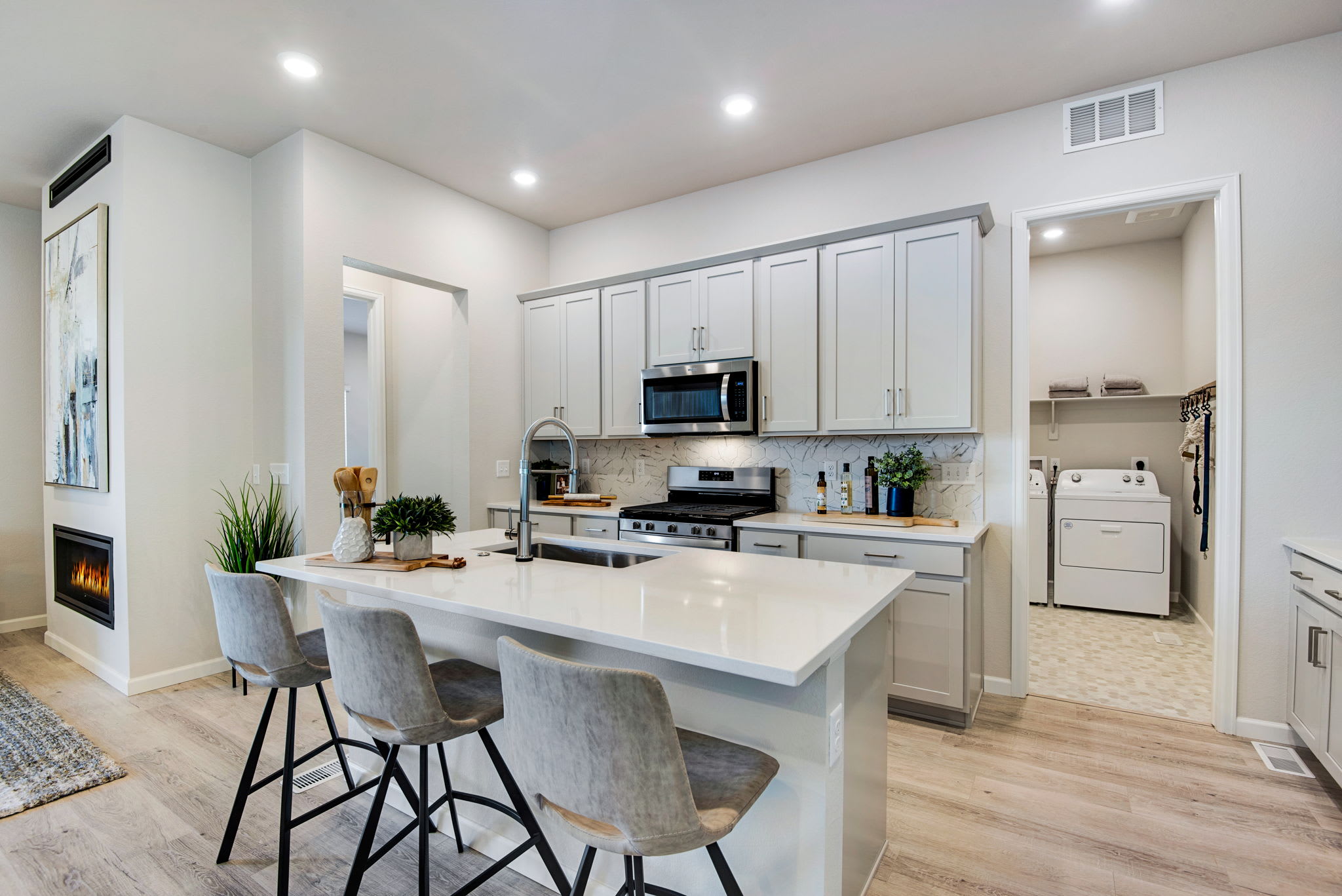 A modern, open-concept kitchen with white cabinets, a large island with bar stools, and hardwood floors, complemented by a fireplace and other home furnishings in the background.