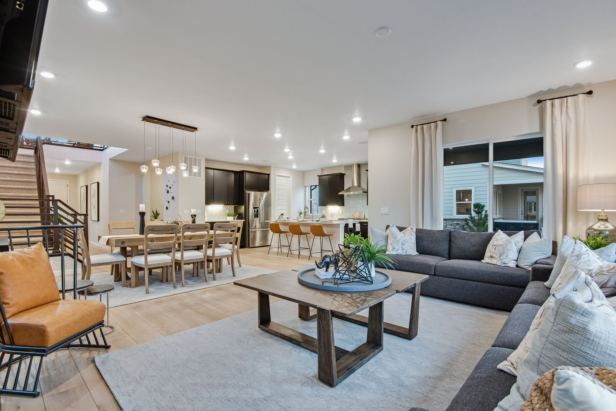 A spacious and modern open-concept living room with a dining area and kitchen, featuring a gray sectional sofa, a wooden coffee table, and various lighting fixtures creating a warm and inviting atmosphere.