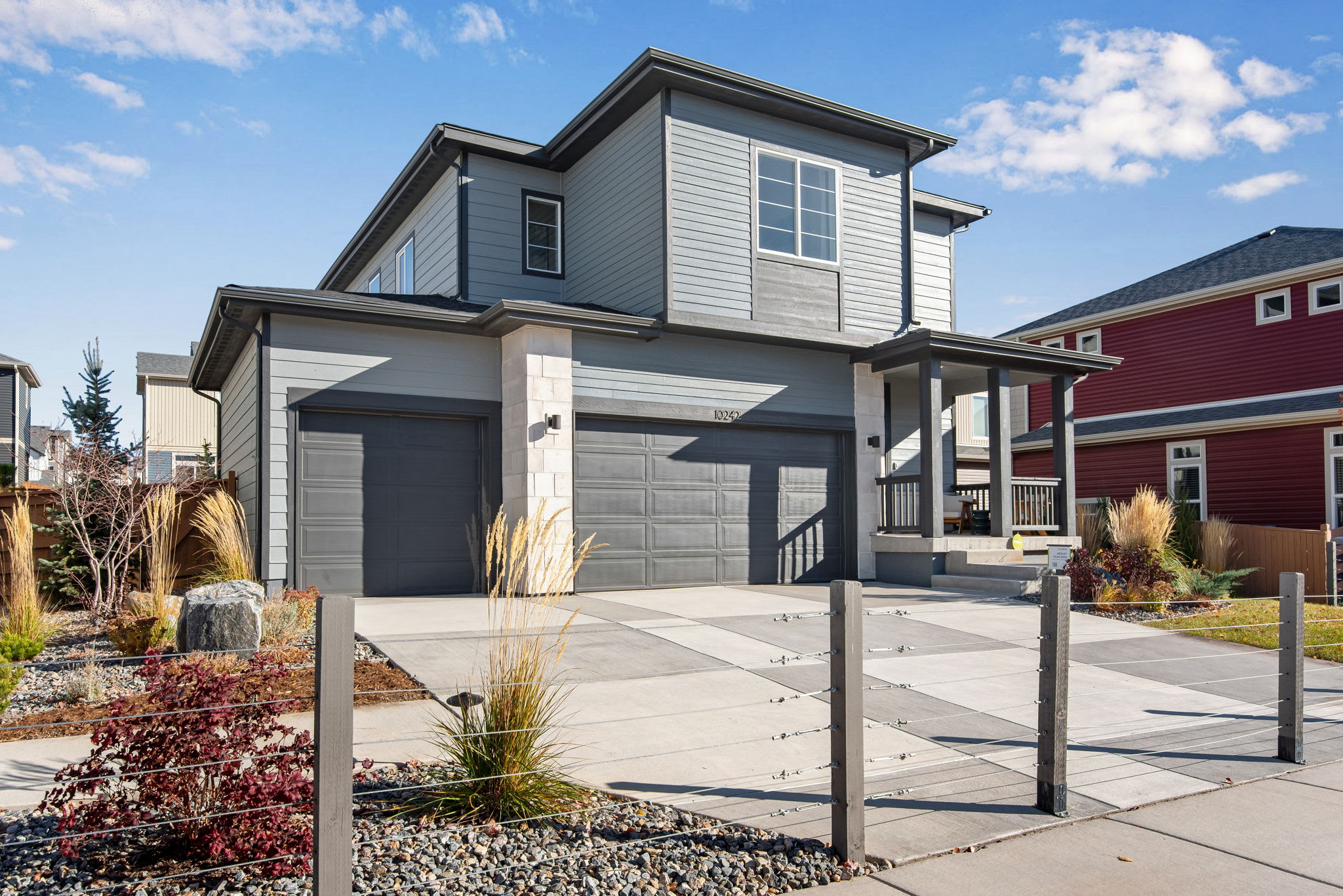 A modern, two-story residential house with a gray exterior, a garage door, and a paved driveway in the foreground, set against a backdrop of a blue sky with scattered clouds.