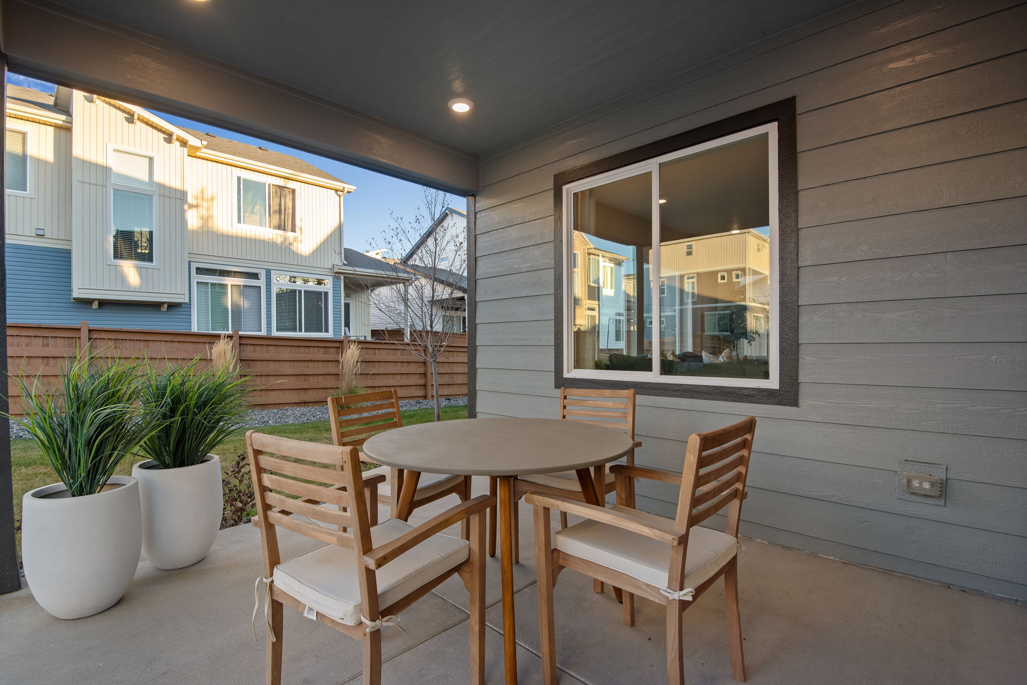 A cozy outdoor patio with a wooden table and chairs, surrounded by potted plants and a modern, gray-toned building in the background.