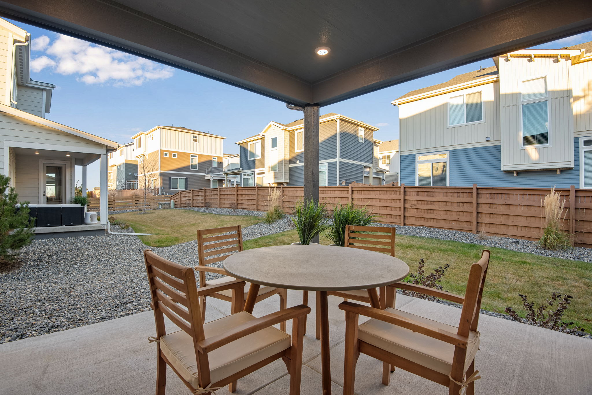 A cozy outdoor seating area with a round wooden table and chairs, surrounded by a gravel patio and fenced-in yards with modern townhouses in the background under a blue sky with clouds.