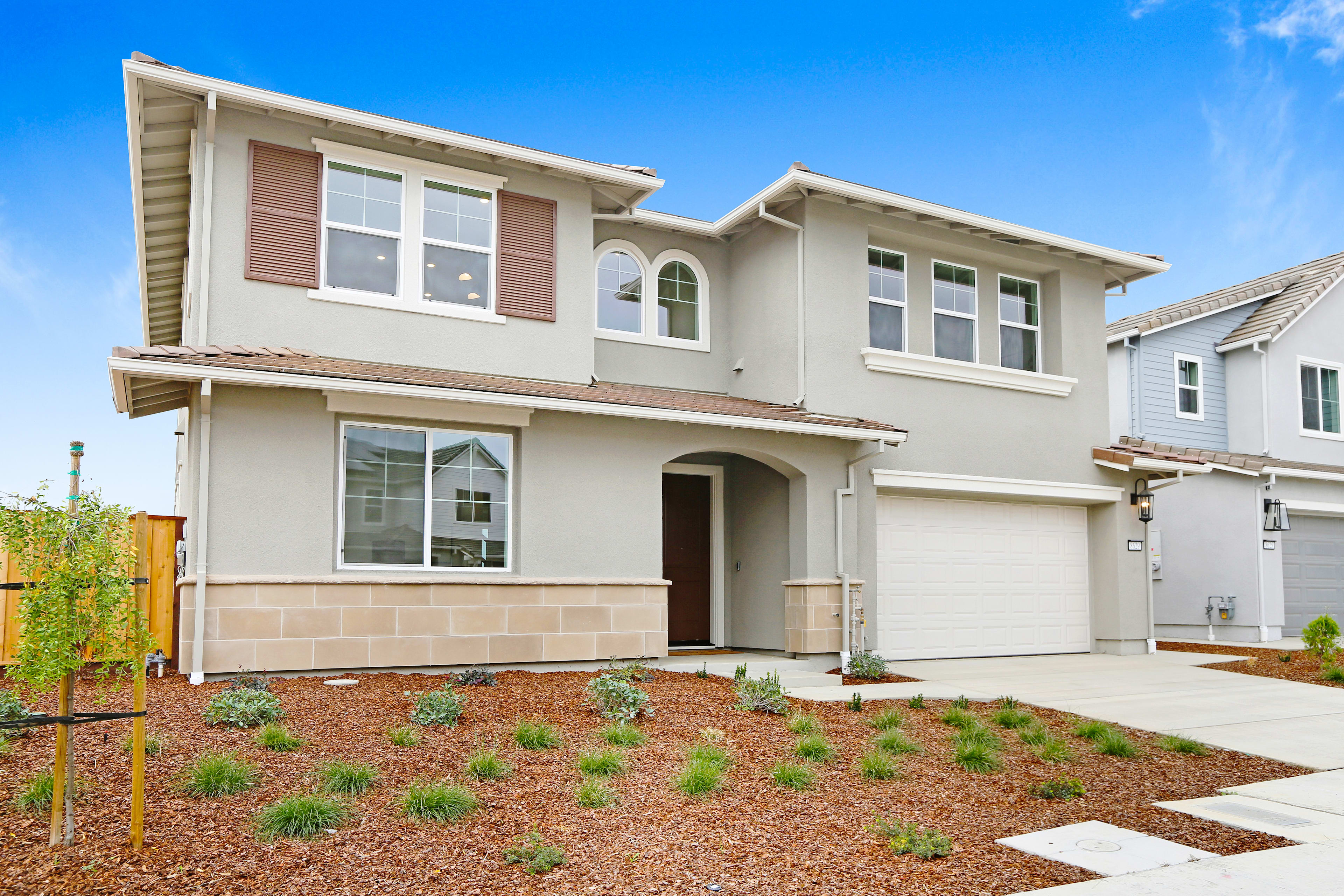 A two-story beige house with a garage, surrounded by a landscaped yard with a grassy area and small plants in the foreground, set against a clear blue sky.