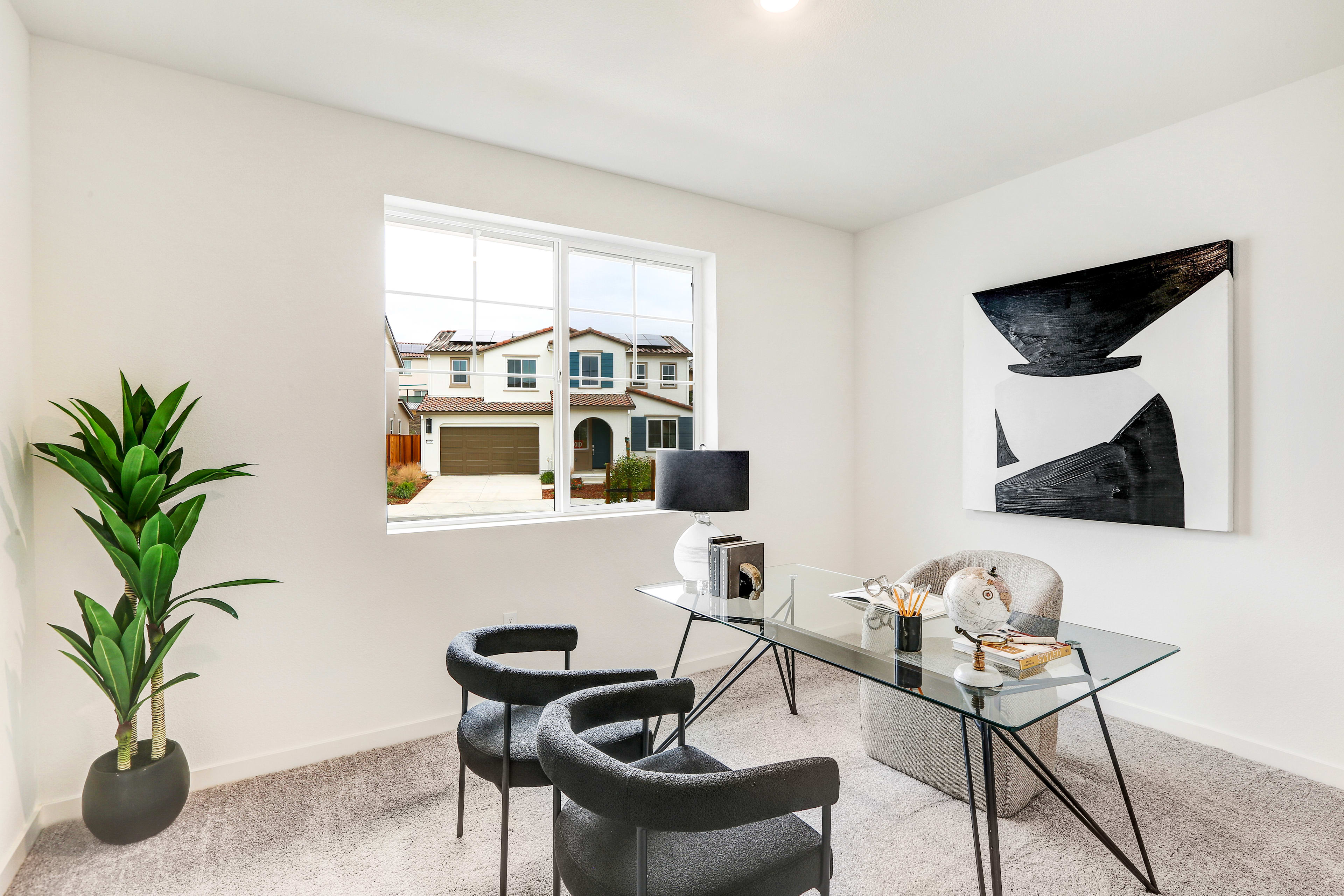A modern and minimalist home office space with a glass desk, black chairs, and abstract artwork on the walls, set against the backdrop of a residential exterior visible through the window.