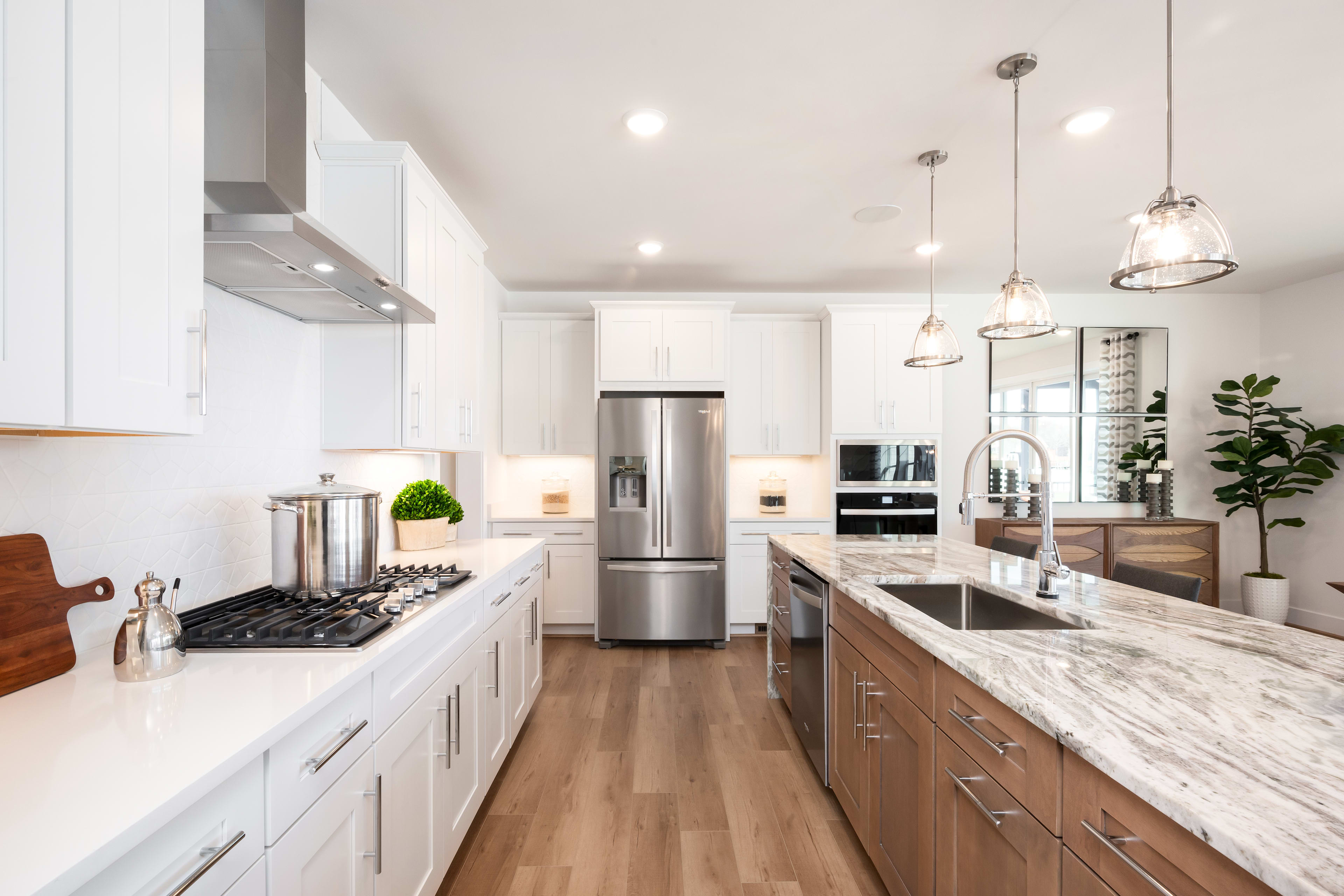 A modern, well-lit kitchen with white cabinets, marble countertops, and stainless steel appliances, featuring a long central island and pendant lighting.