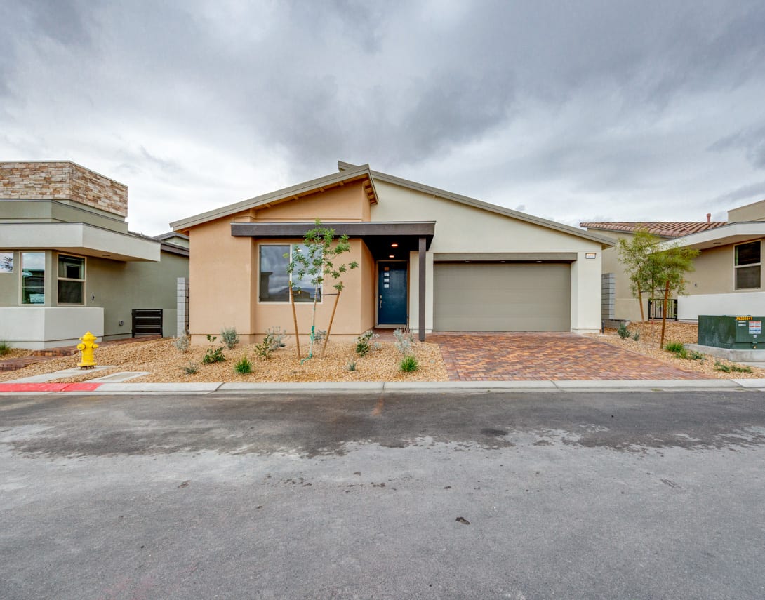 A single-story residential home with a brick and stucco exterior, surrounded by a paved driveway and landscaping, set against a cloudy sky.