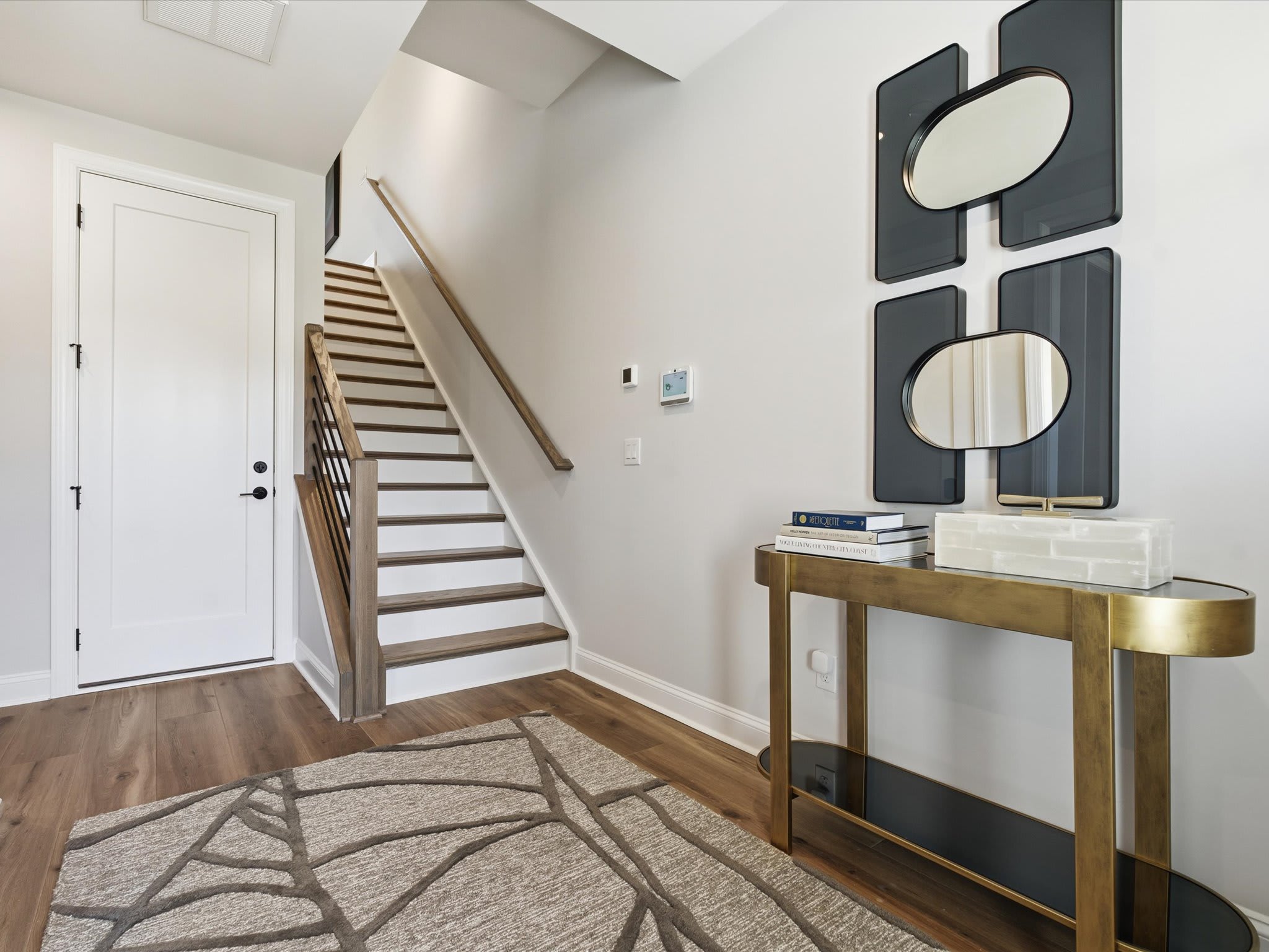 A modern and minimalist entryway with a wooden staircase, a console table with decorative mirrors, and a patterned area rug on the hardwood floor.