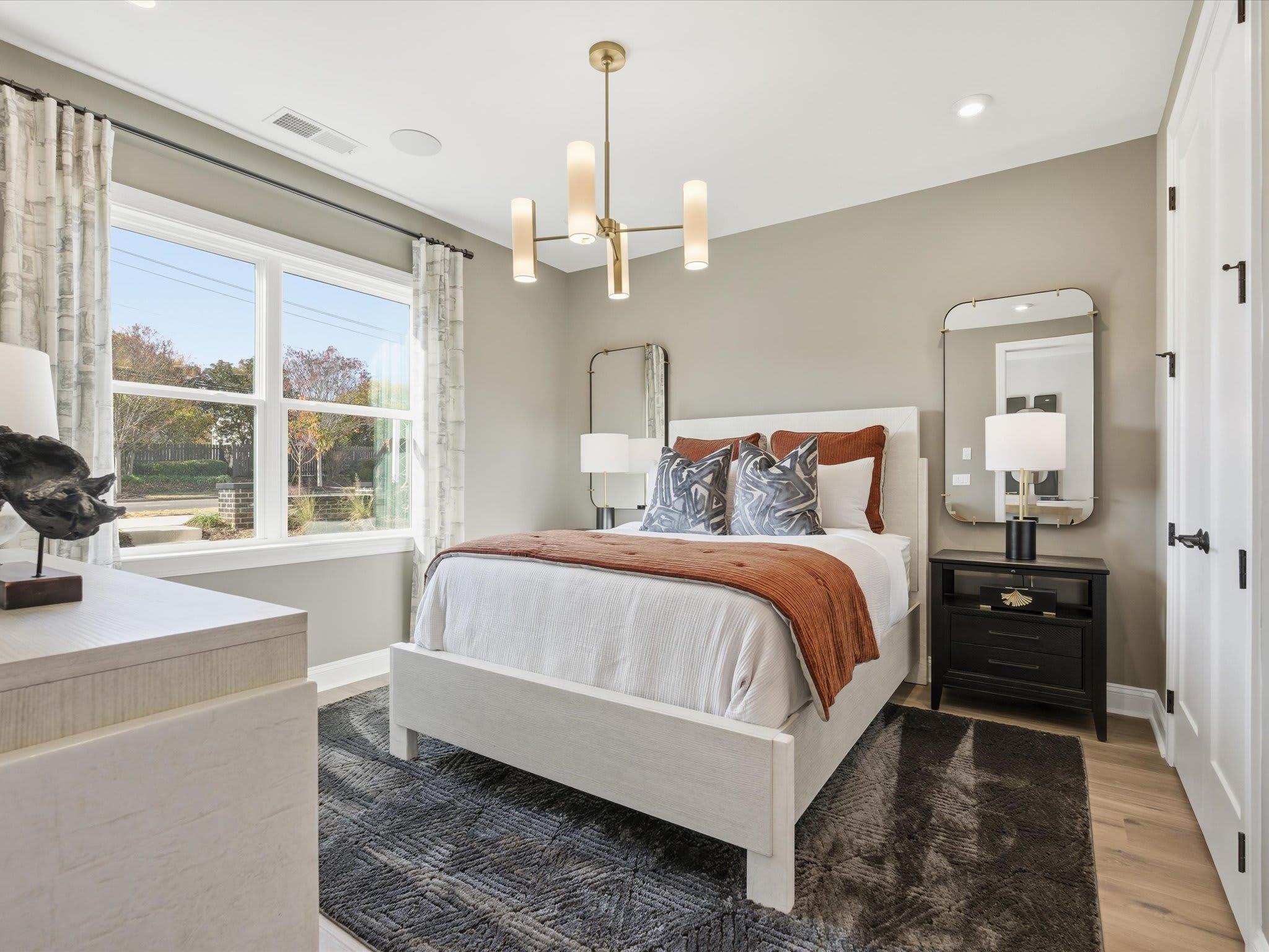 A cozy and well-appointed bedroom with a large window, a wooden bed frame, and a modern chandelier overhead, set against a neutral color palette and accented with textured bedding and decor.