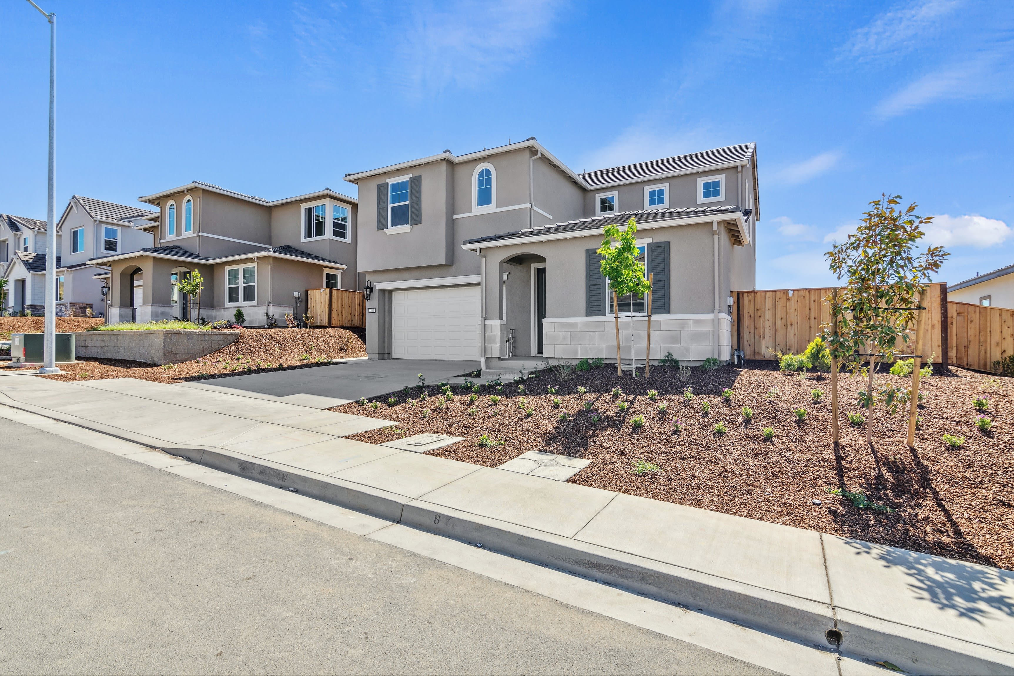 A newly constructed two-story residential home with a landscaped front yard, surrounded by other similar houses in a suburban neighborhood under a clear blue sky.