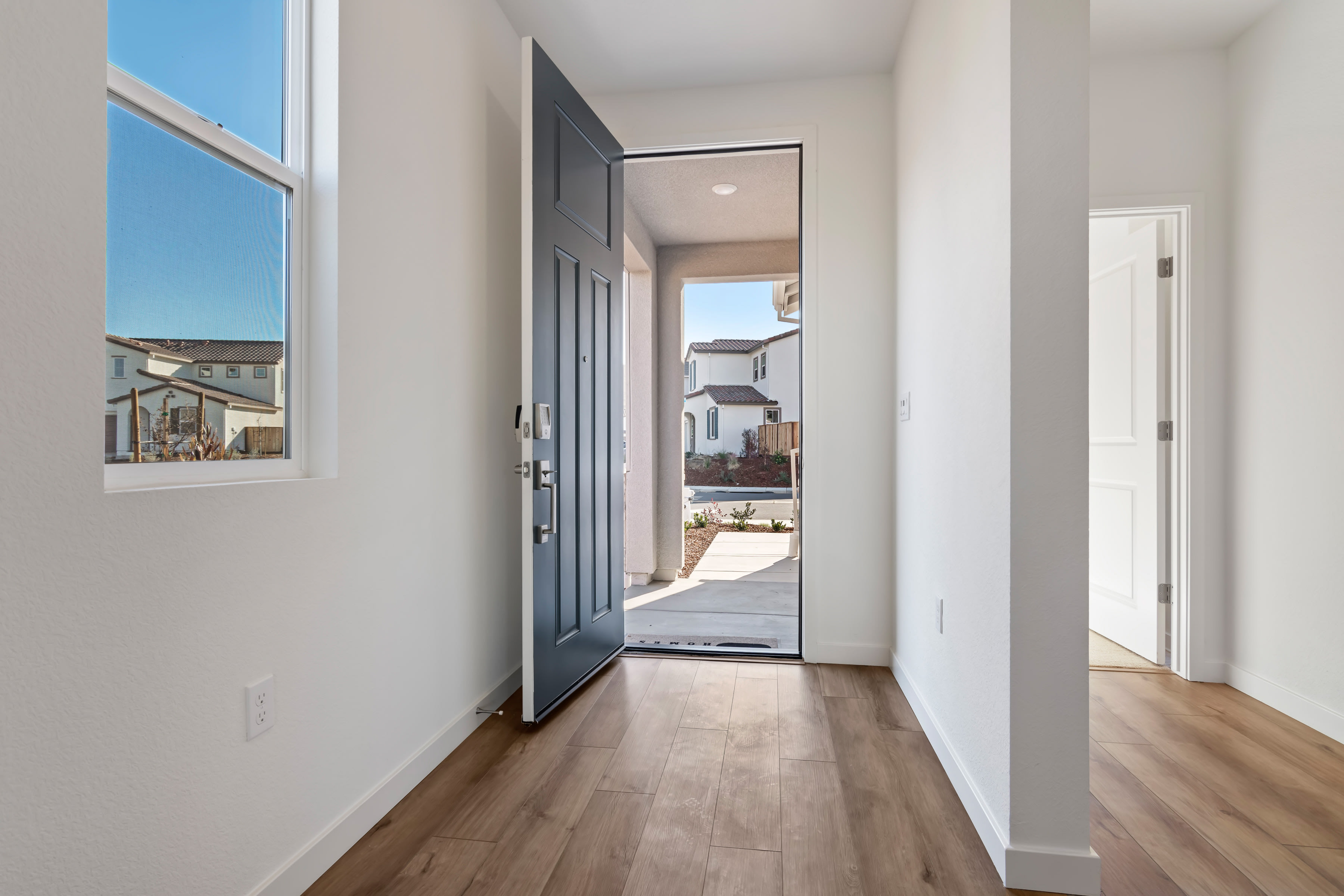 A bright and airy hallway with a wooden floor, leading to an open doorway that reveals a view of the outdoors.