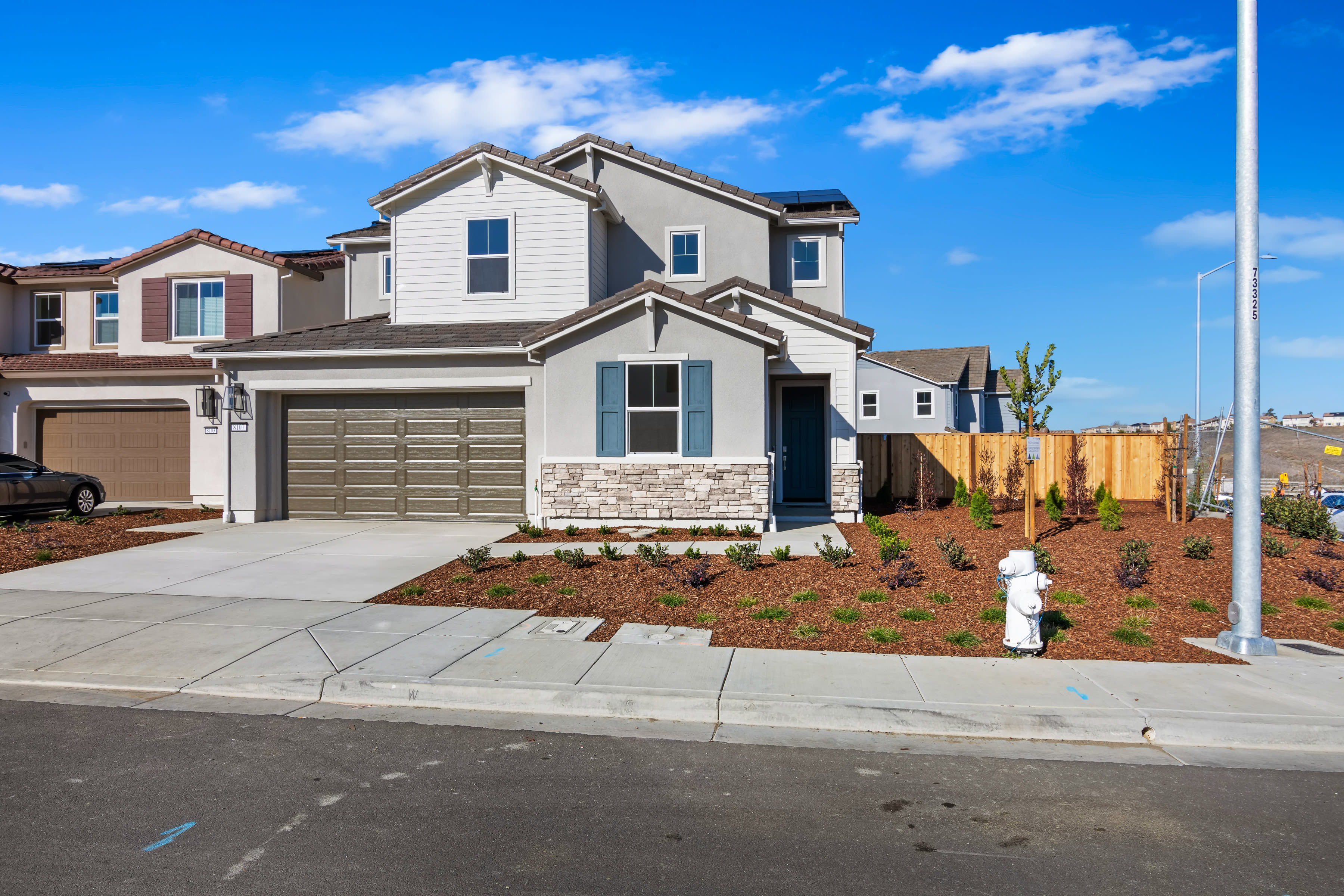A two-story residential house with a garage, surrounded by a landscaped yard and set against a clear blue sky with scattered clouds.