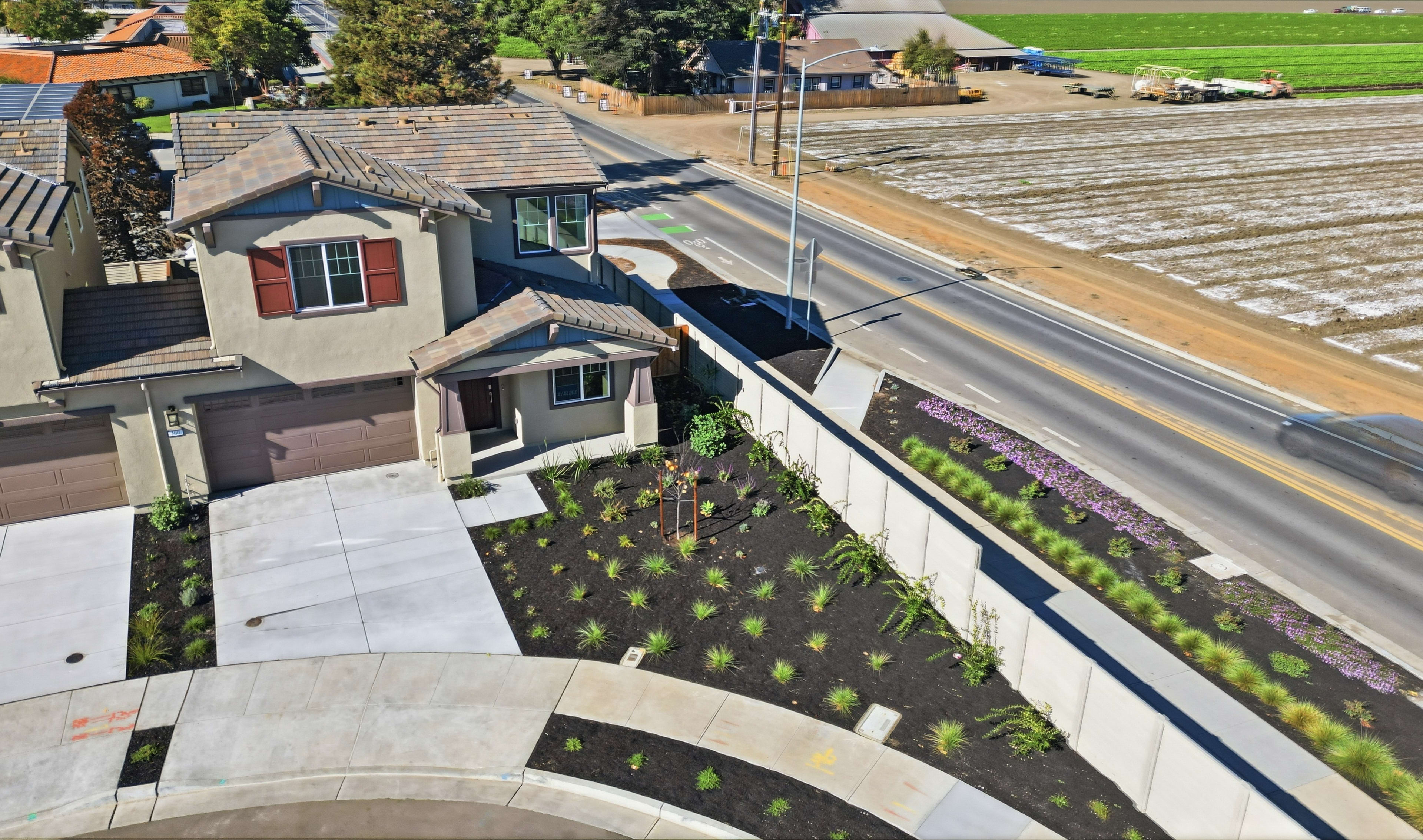 A residential neighborhood with a winding path, landscaped gardens, and a view of agricultural fields in the background.