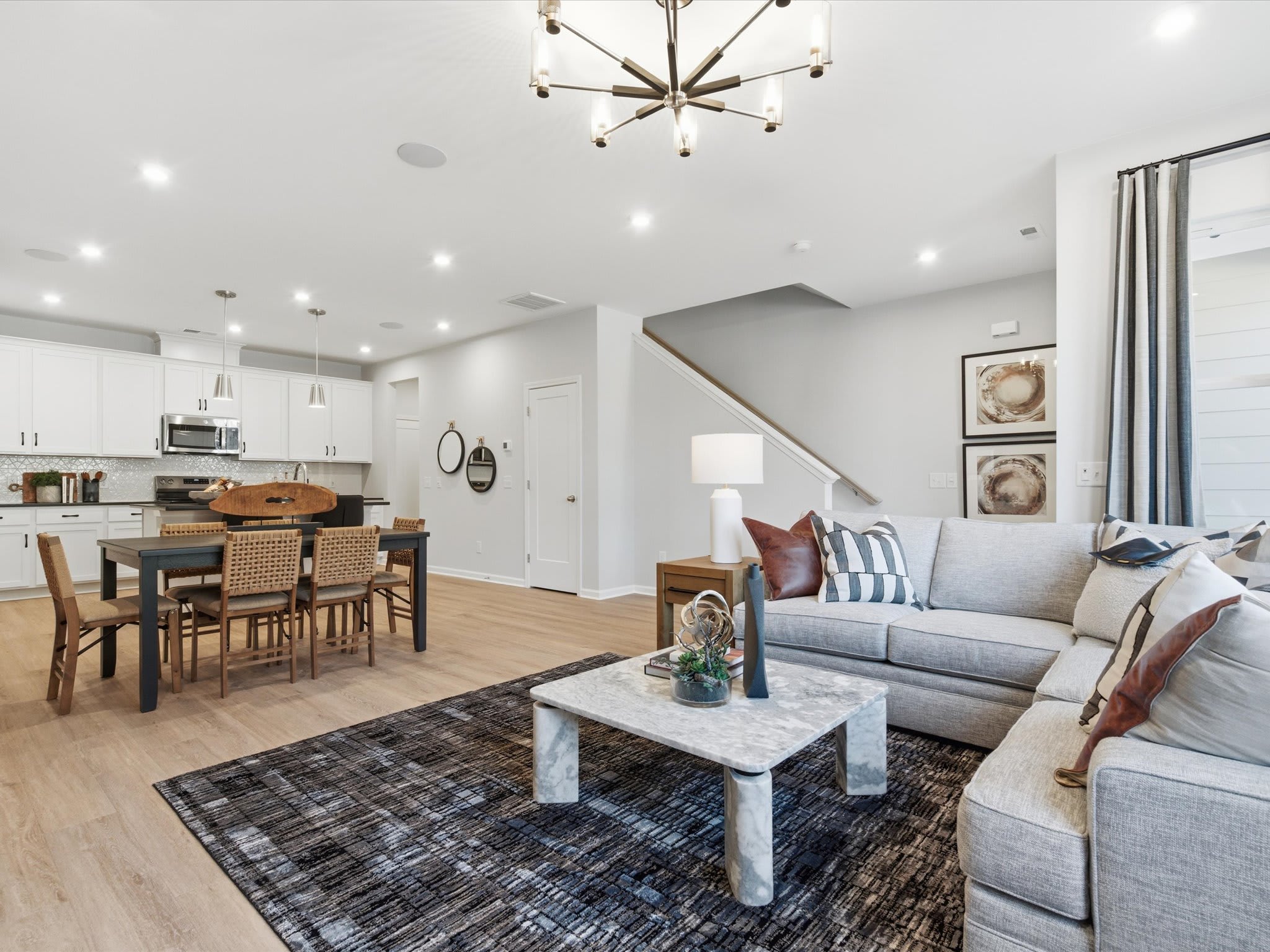 A modern and spacious open-concept living room with a dining area and kitchen, featuring a large gray sofa, a marble coffee table, and a statement light fixture overhead.