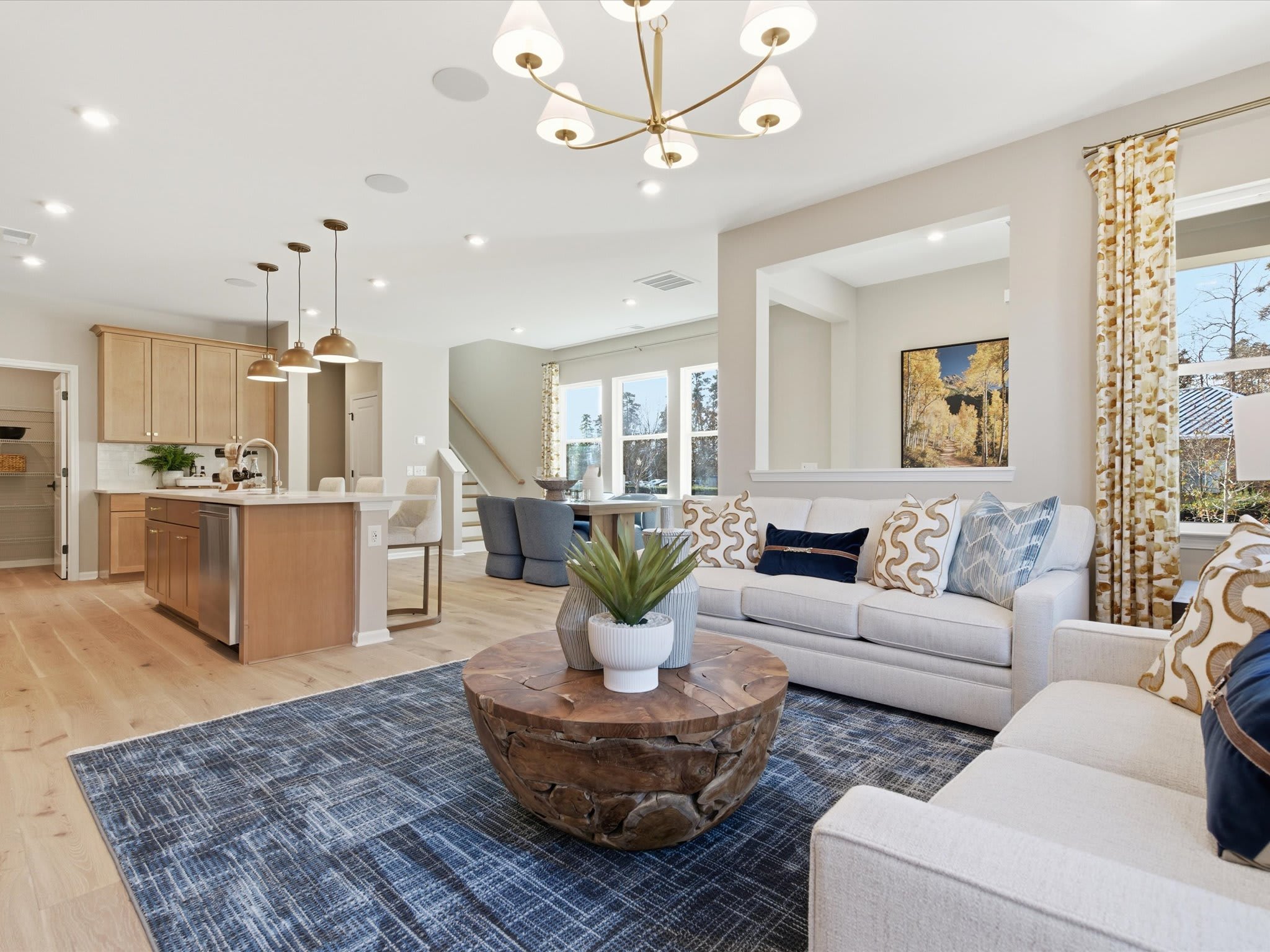 A cozy and well-furnished living room with a modern chandelier, a wooden coffee table, and a comfortable sofa set against a backdrop of a kitchen area with stainless steel appliances.