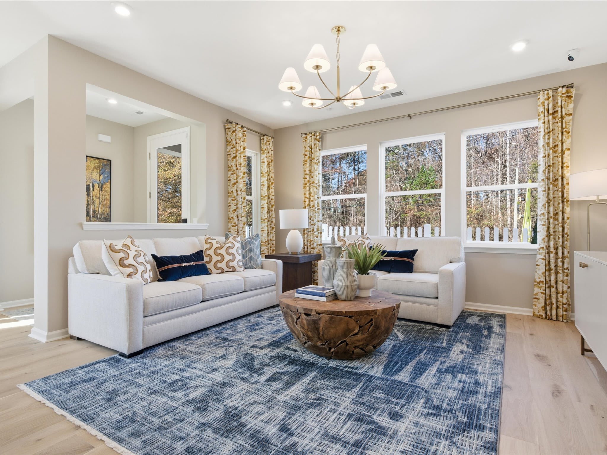 A cozy and well-furnished living room with a plush sofa, armchairs, a patterned rug, and a chandelier overhead, surrounded by large windows that provide a view of the outdoor scenery.