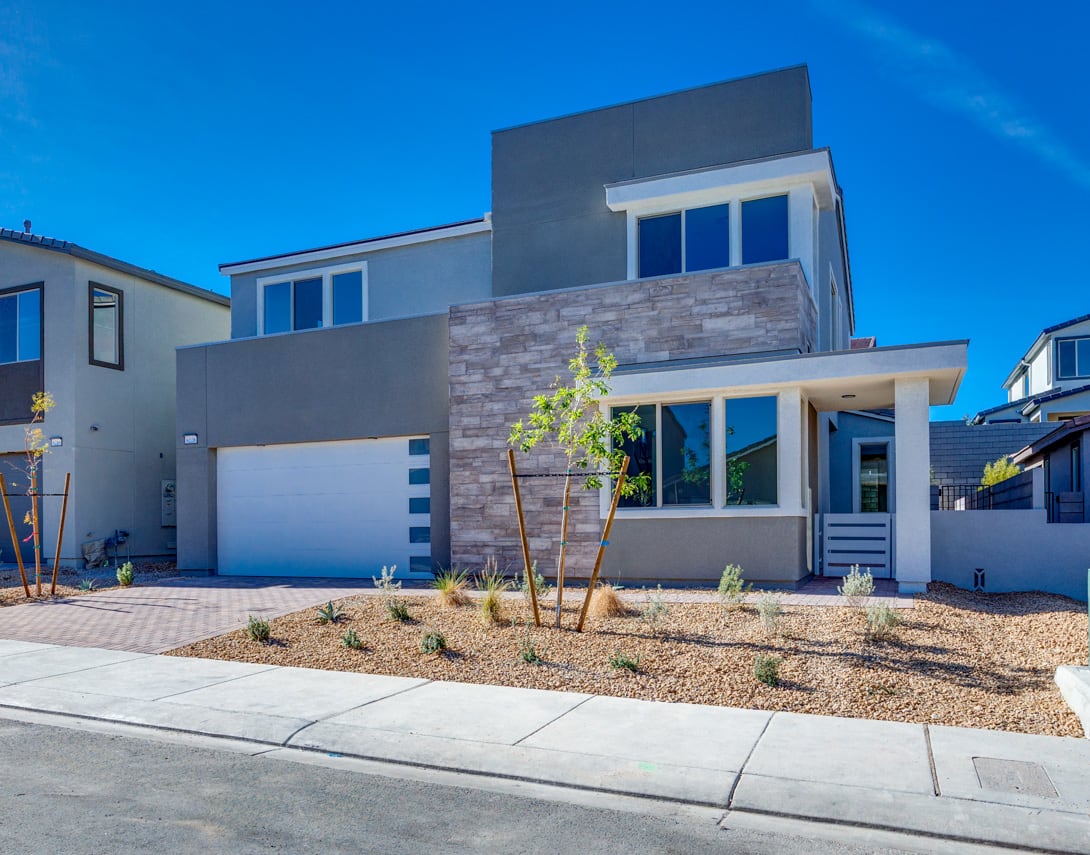 A modern, two-story residential building with a gray exterior, large windows, and a landscaped front yard featuring desert-adapted plants and a paved driveway, set against a clear blue sky.