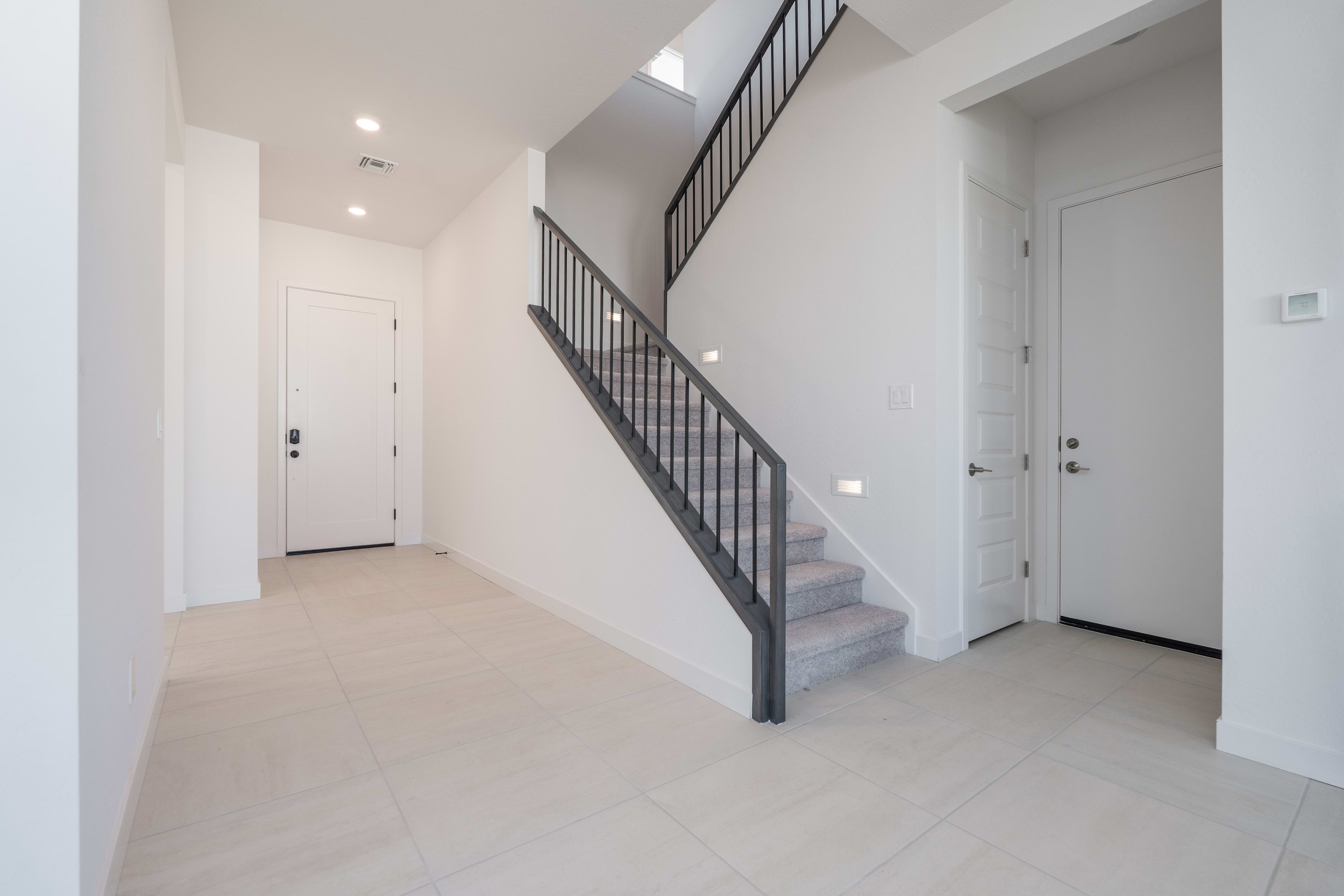 A bright, minimalist hallway with a staircase leading upstairs, featuring white walls, a tiled floor, and a black metal railing.