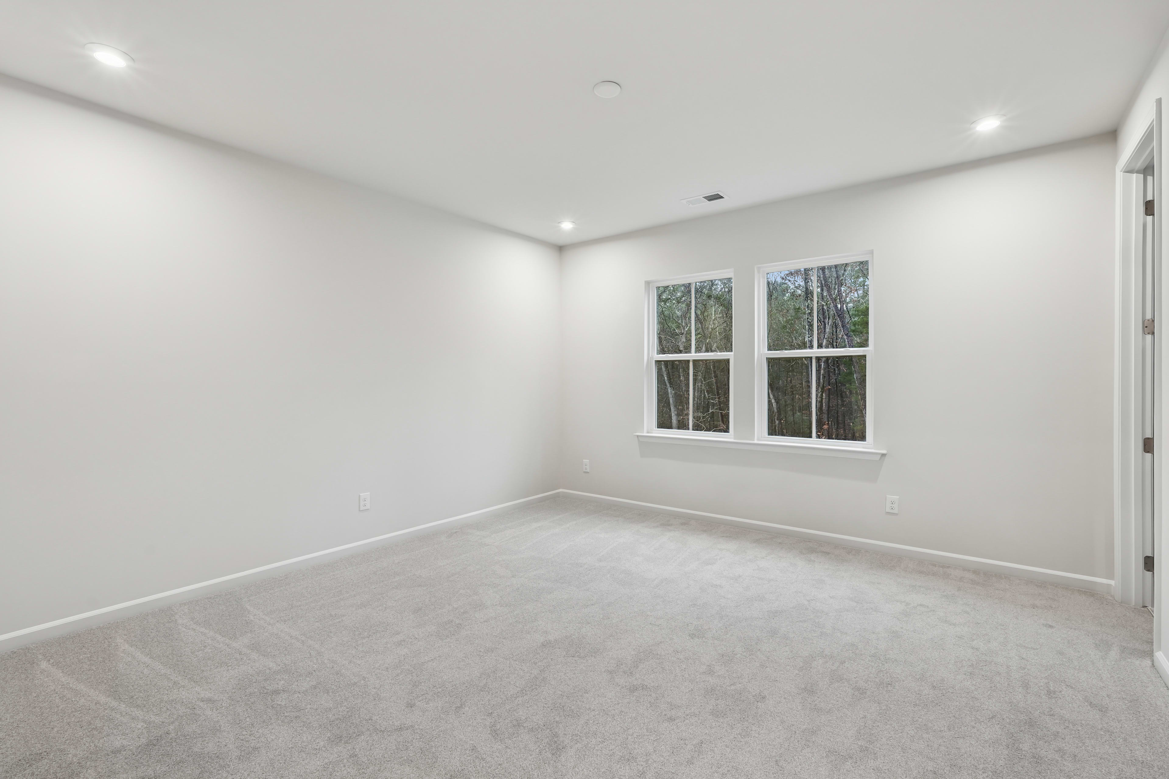 A spacious, well-lit room with white walls, a carpeted floor, and large windows overlooking the outdoors.