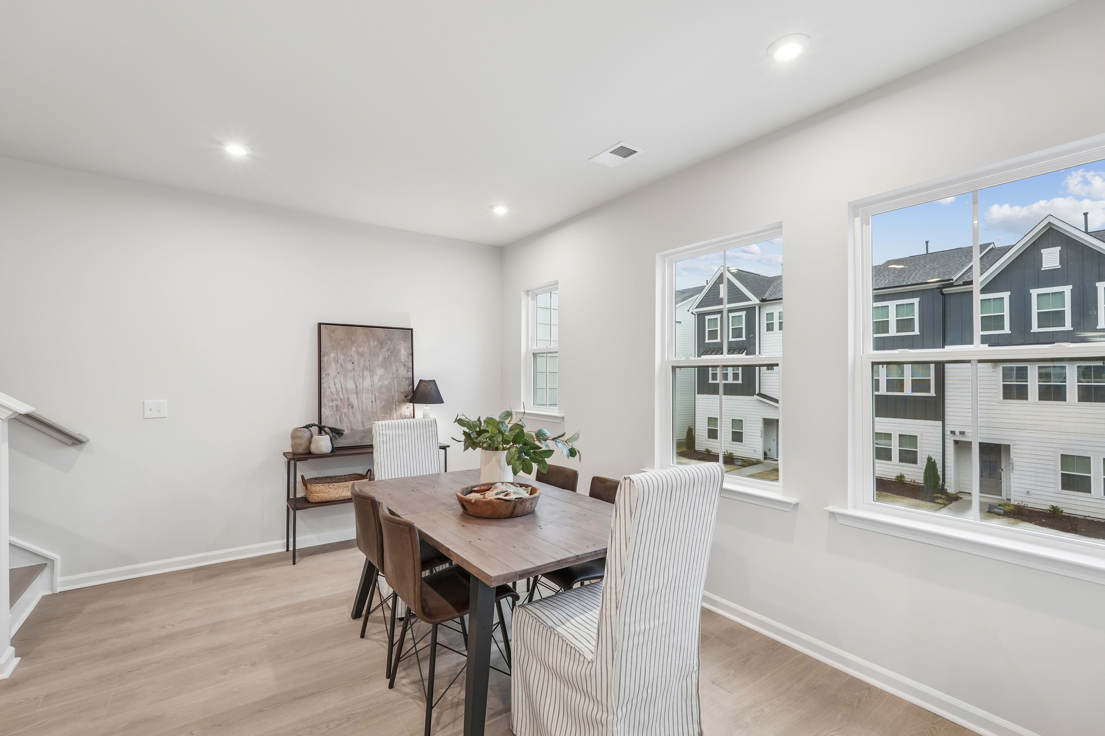 A modern, open-concept dining area with a wooden table and chairs, surrounded by large windows overlooking a residential neighborhood.